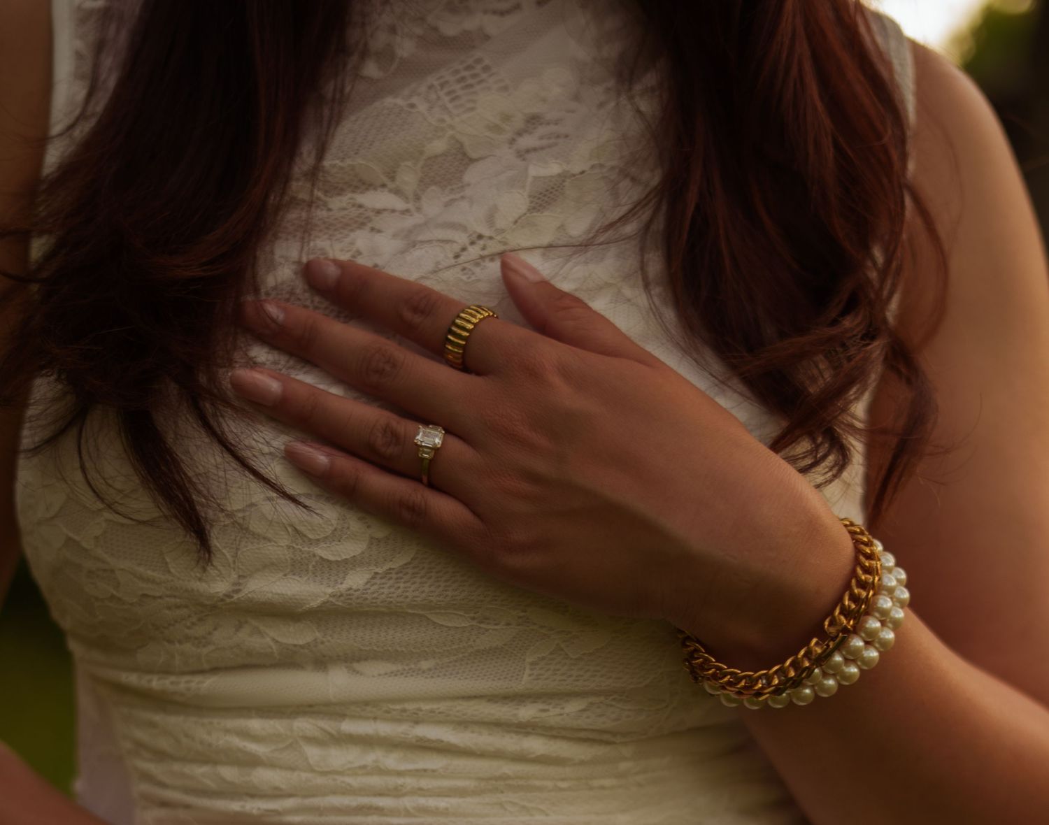 Woman in white lace dress wearing gold rings and pearl bracelet with hand placed on chest.
