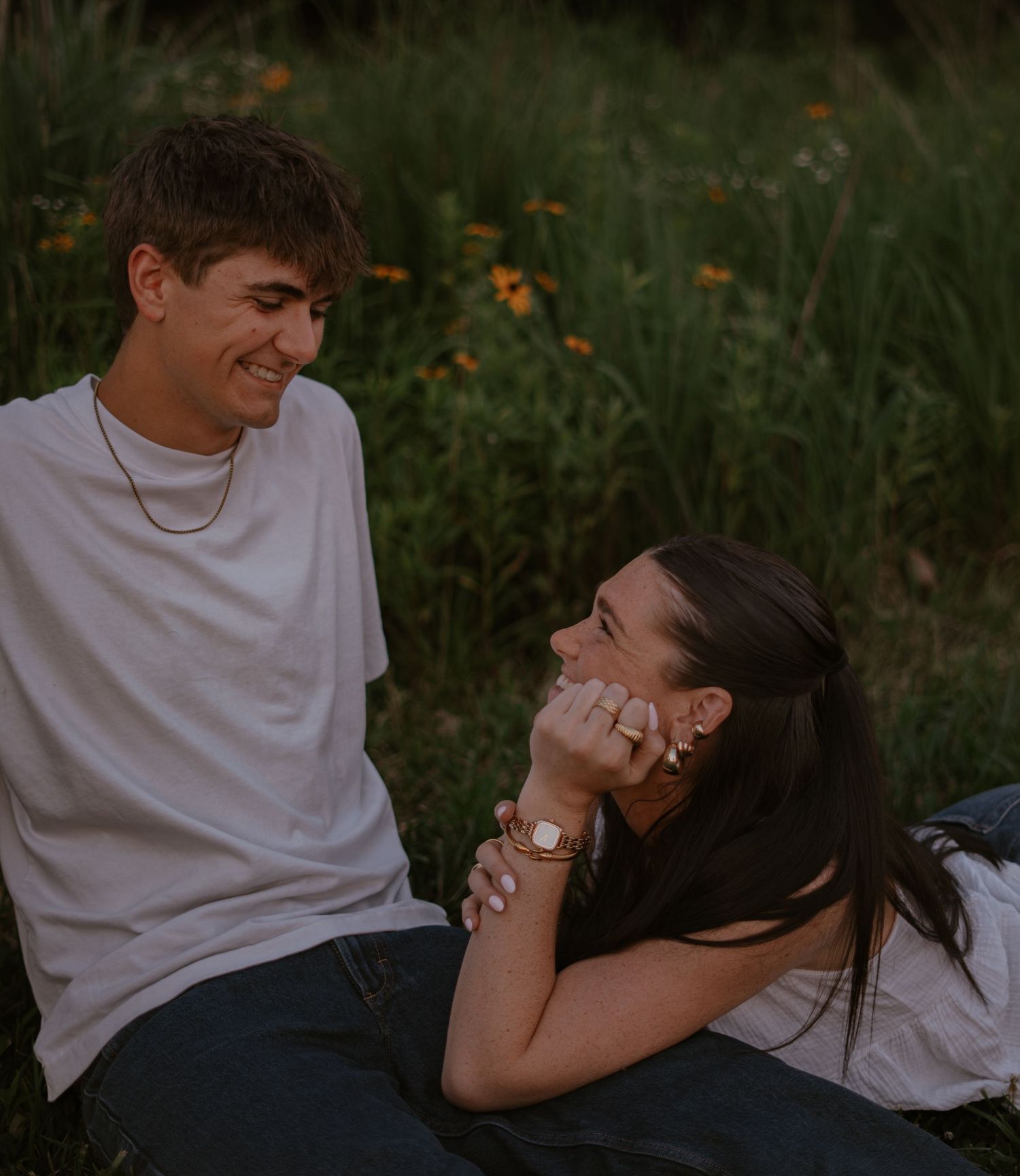 A young couple sits together in a lush green field, smiling and laughing during a romantic outdoor photo session.