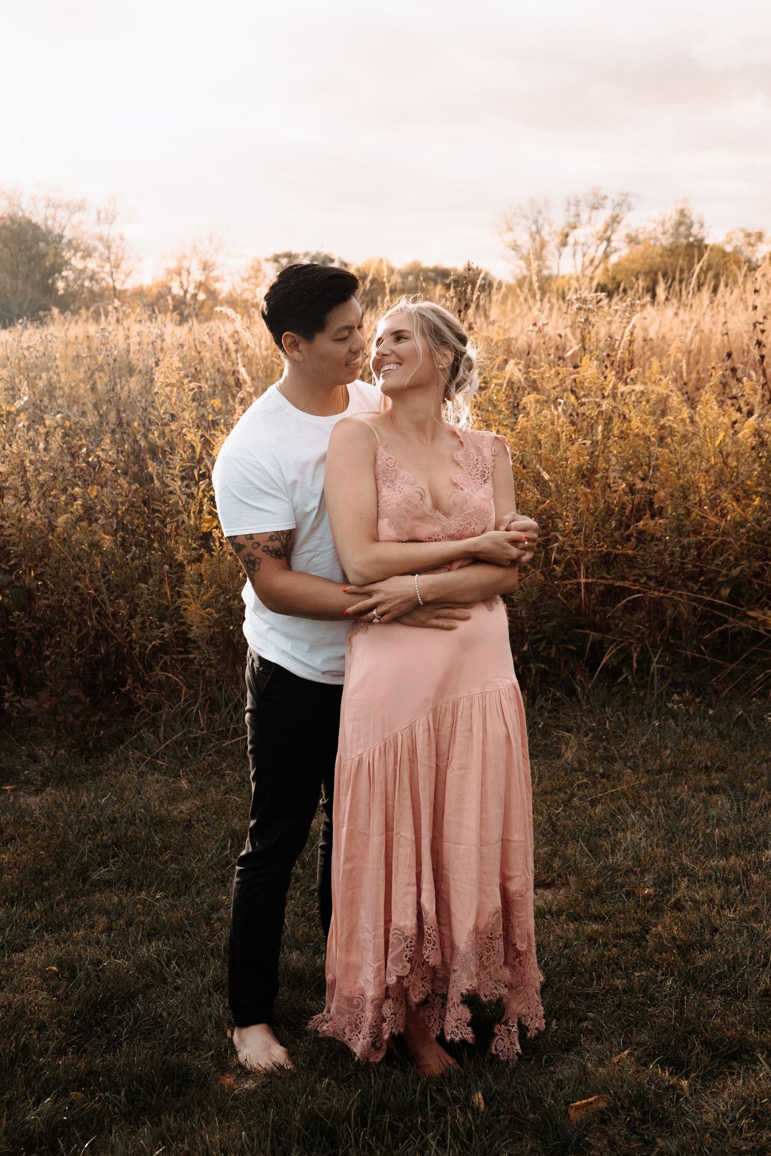 A couple embraces lovingly in a golden field at sunset while wearing casual attire.