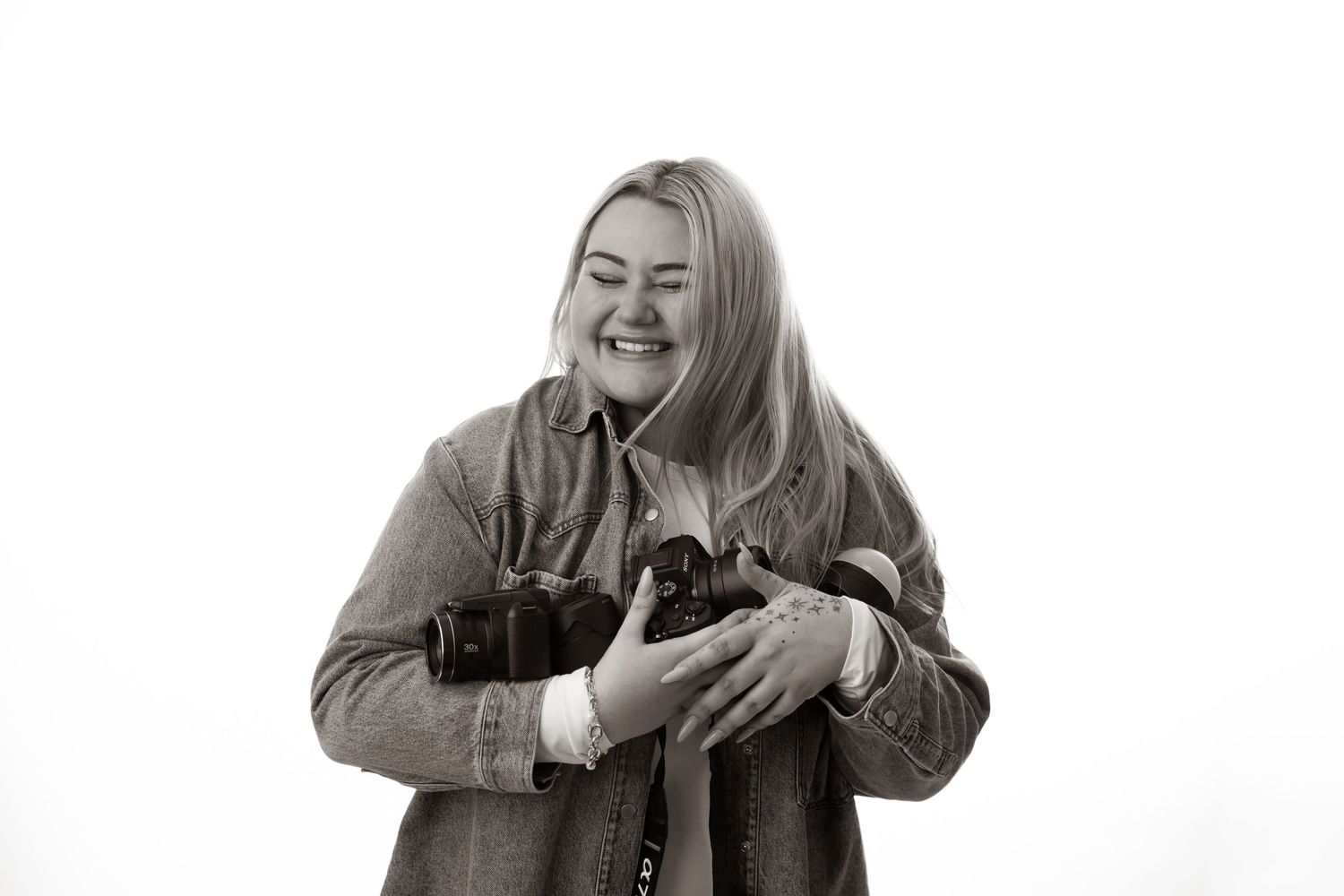 Black and white photo of a smiling blonde woman holding two cameras against a white background.