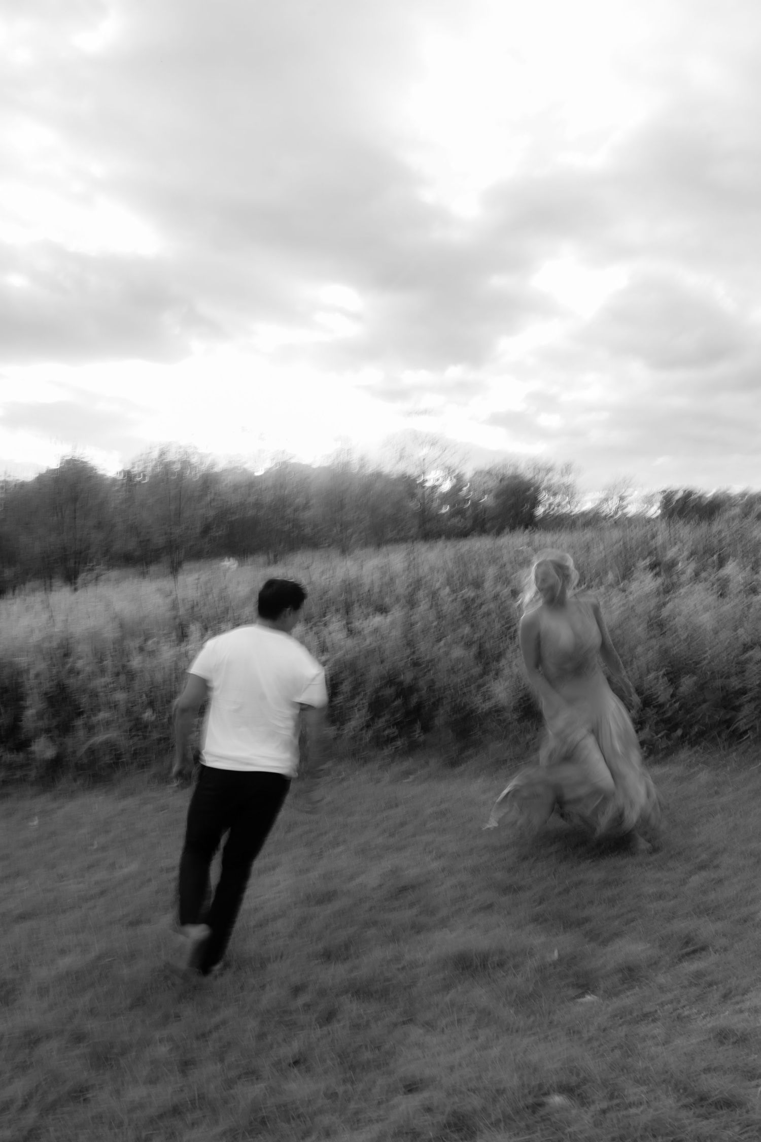 A blurry black and white photo shows two people running through a grassy field under cloudy skies.
