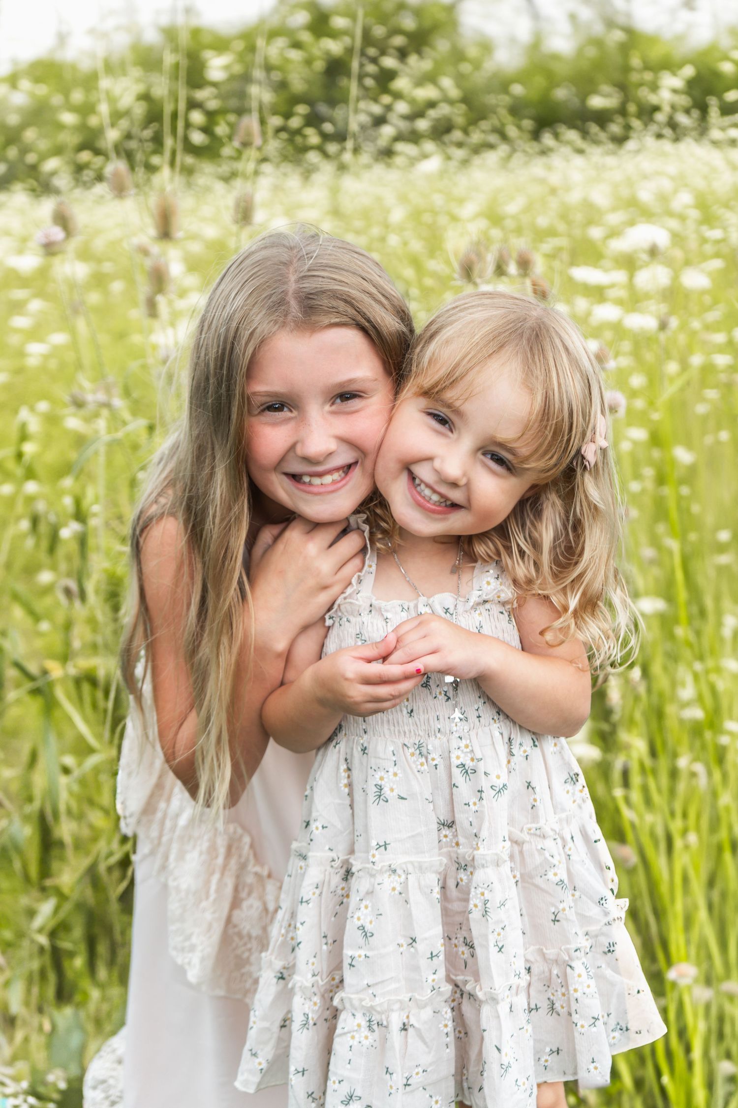 Two children in white summer dresses share a joyful moment together in a sunny meadow filled with wildflowers.