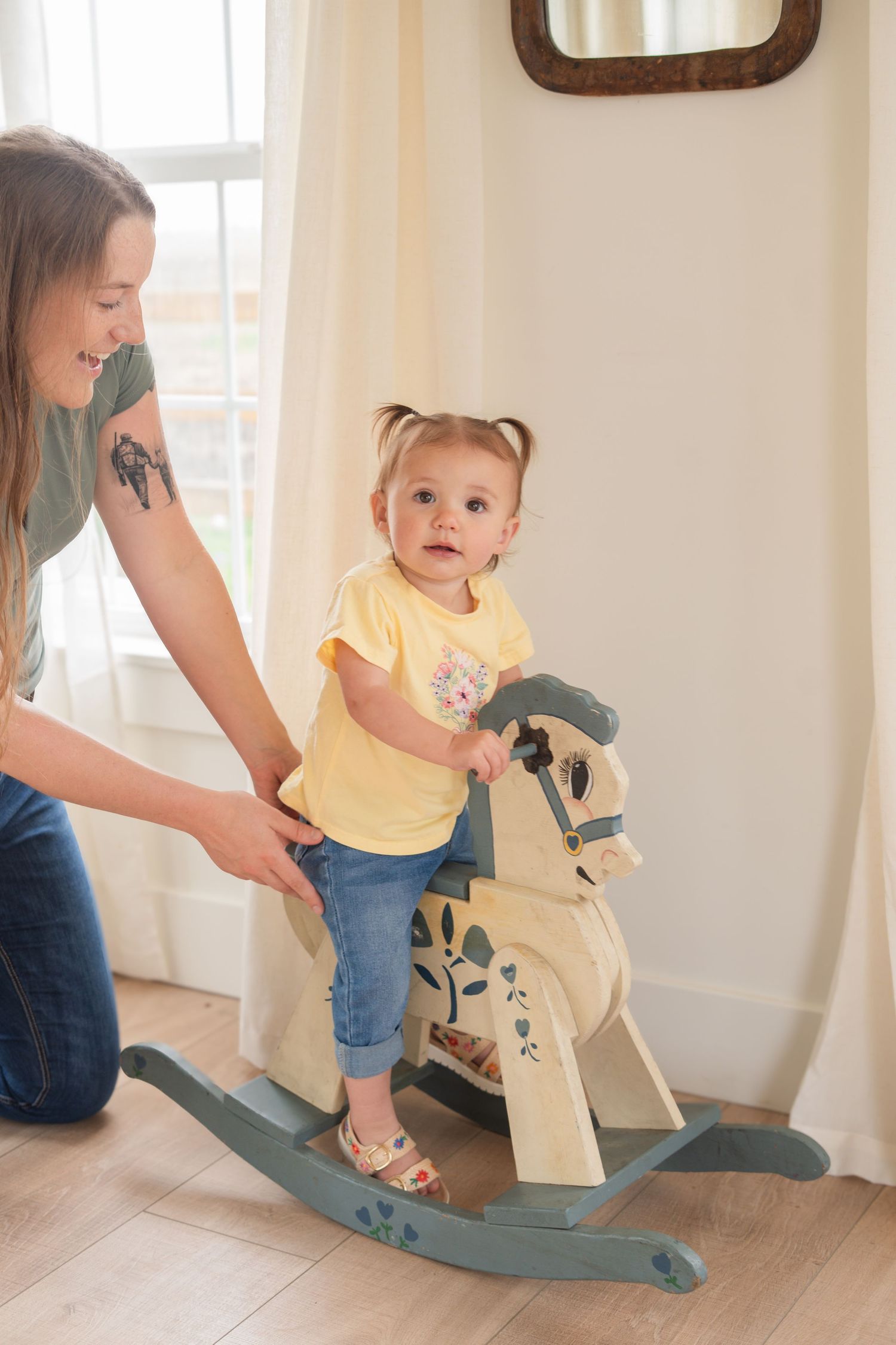 Mother helping toddler girl ride a wooden rocking horse in a bright living room.