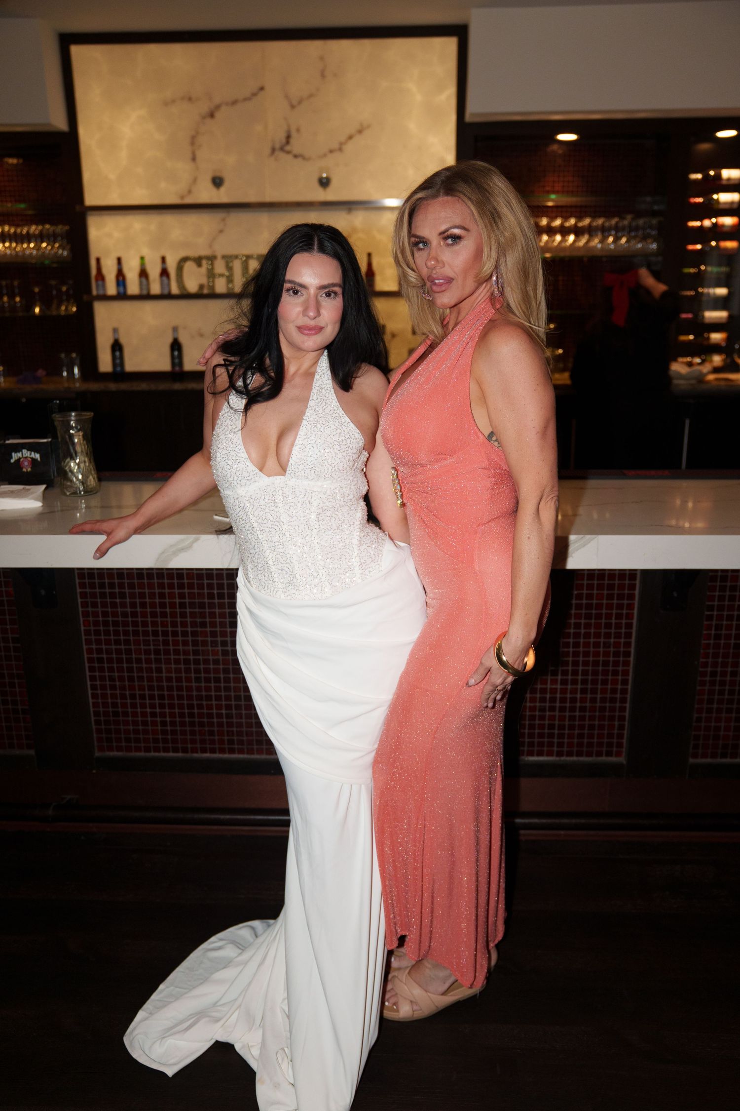 Two elegantly dressed women pose together at a bar, one in a white bridal gown and one in a coral halter dress.