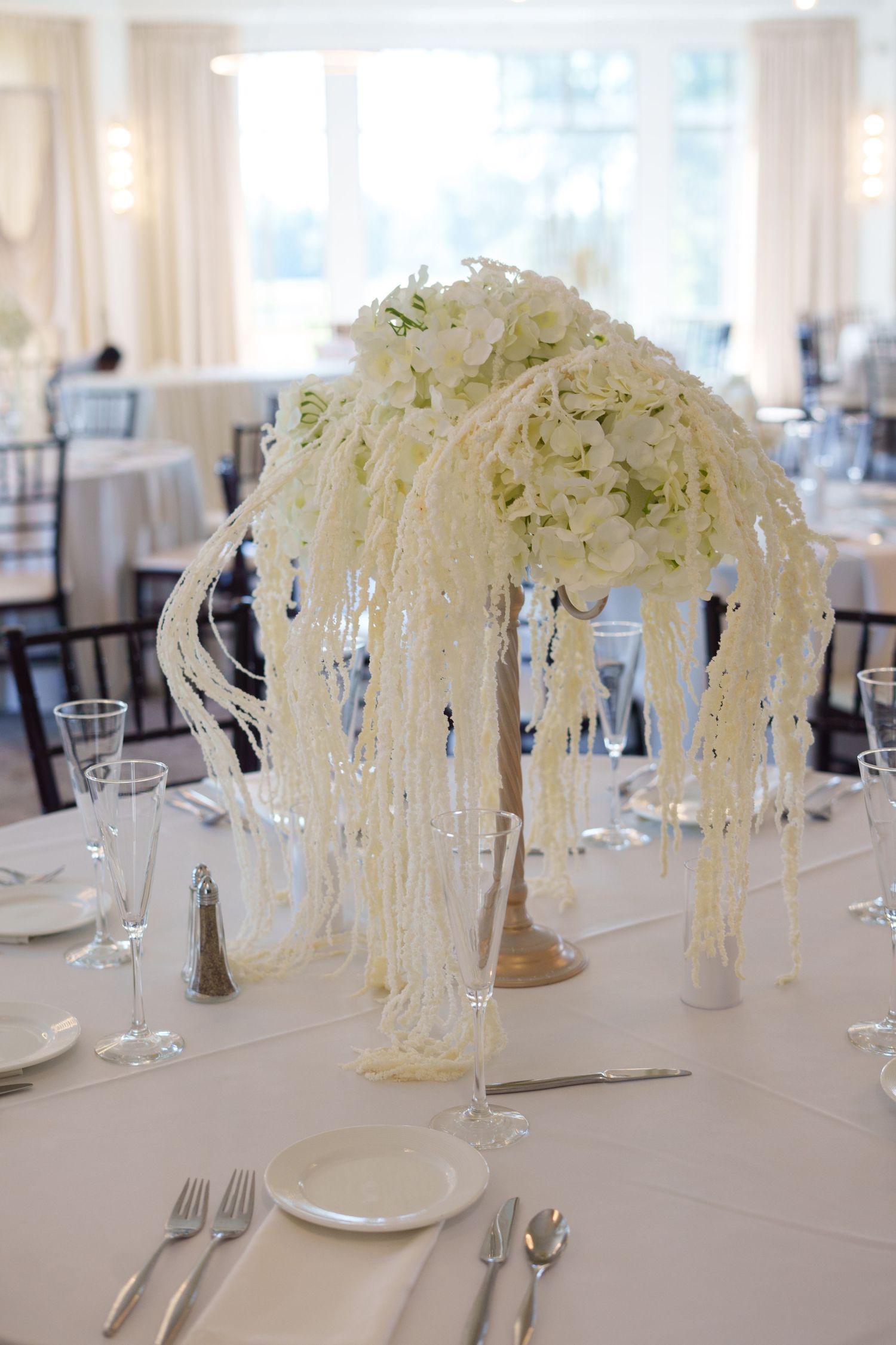 Tall white floral centerpiece with cascading pearl strands on white linen table setting at wedding reception.