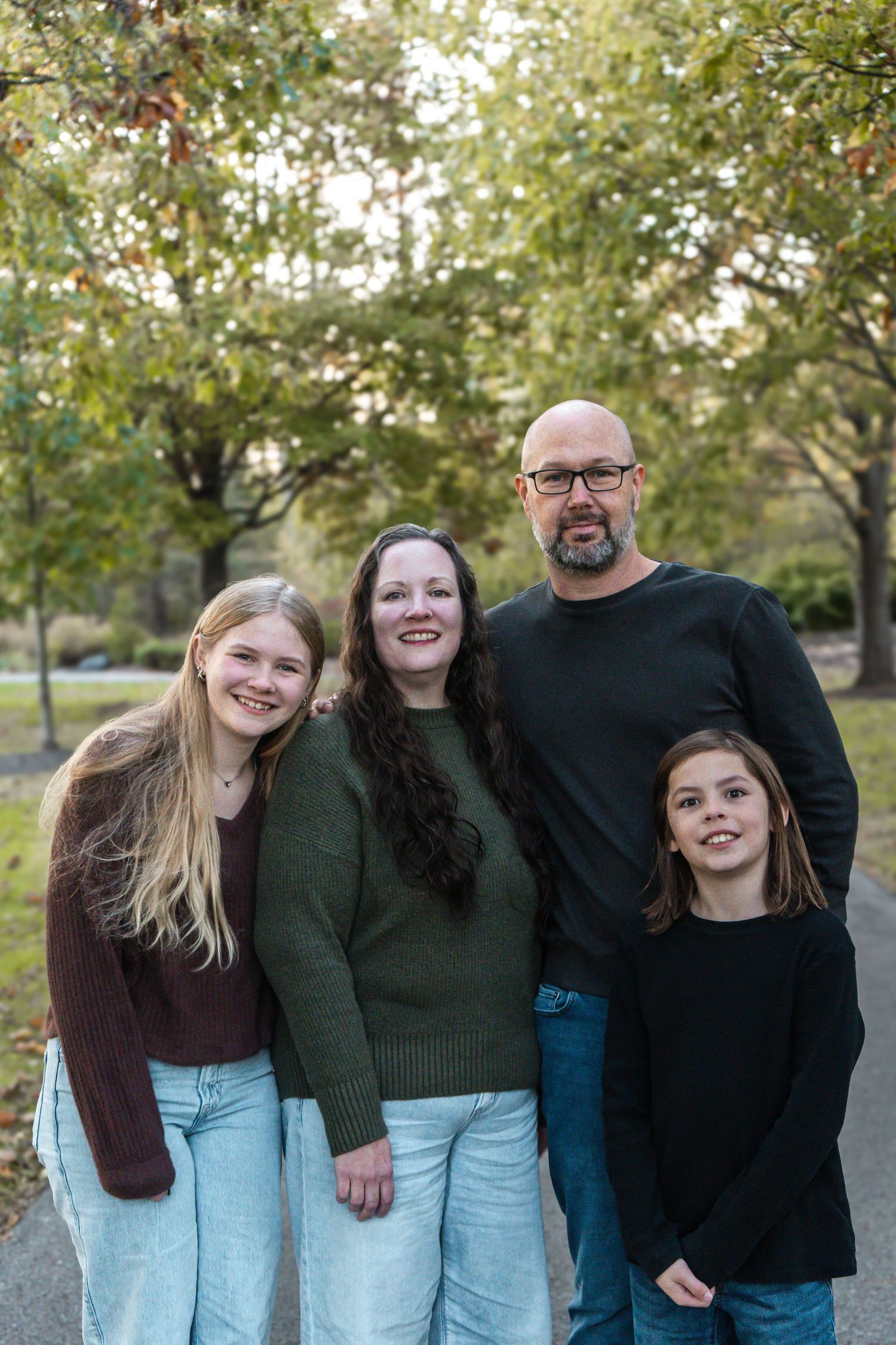 A family wearing coordinated dark sweaters and jeans poses together outdoors in an autumn setting.