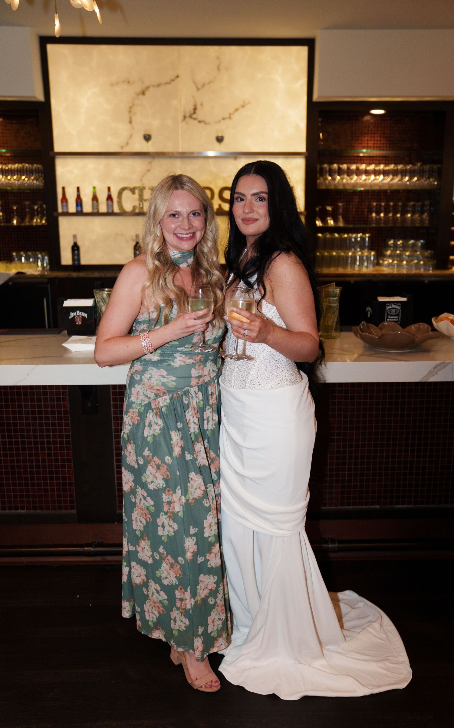 Two women posing at a bar, one in a floral maxi dress and one in a white bridal gown, holding champagne glasses.