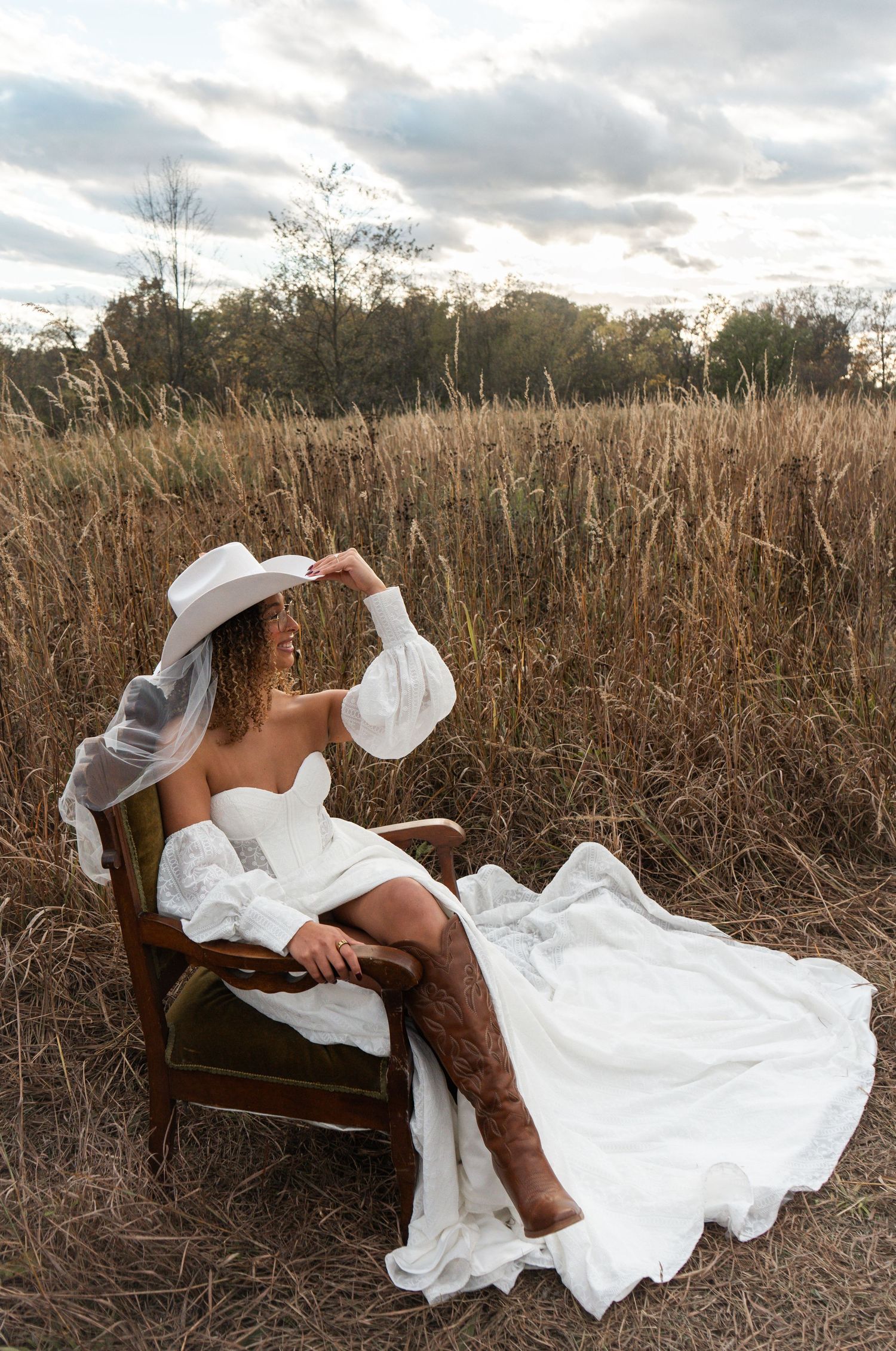 Bride in white mini dress, cowboy hat, and brown boots sits in vintage chair among golden field grass.