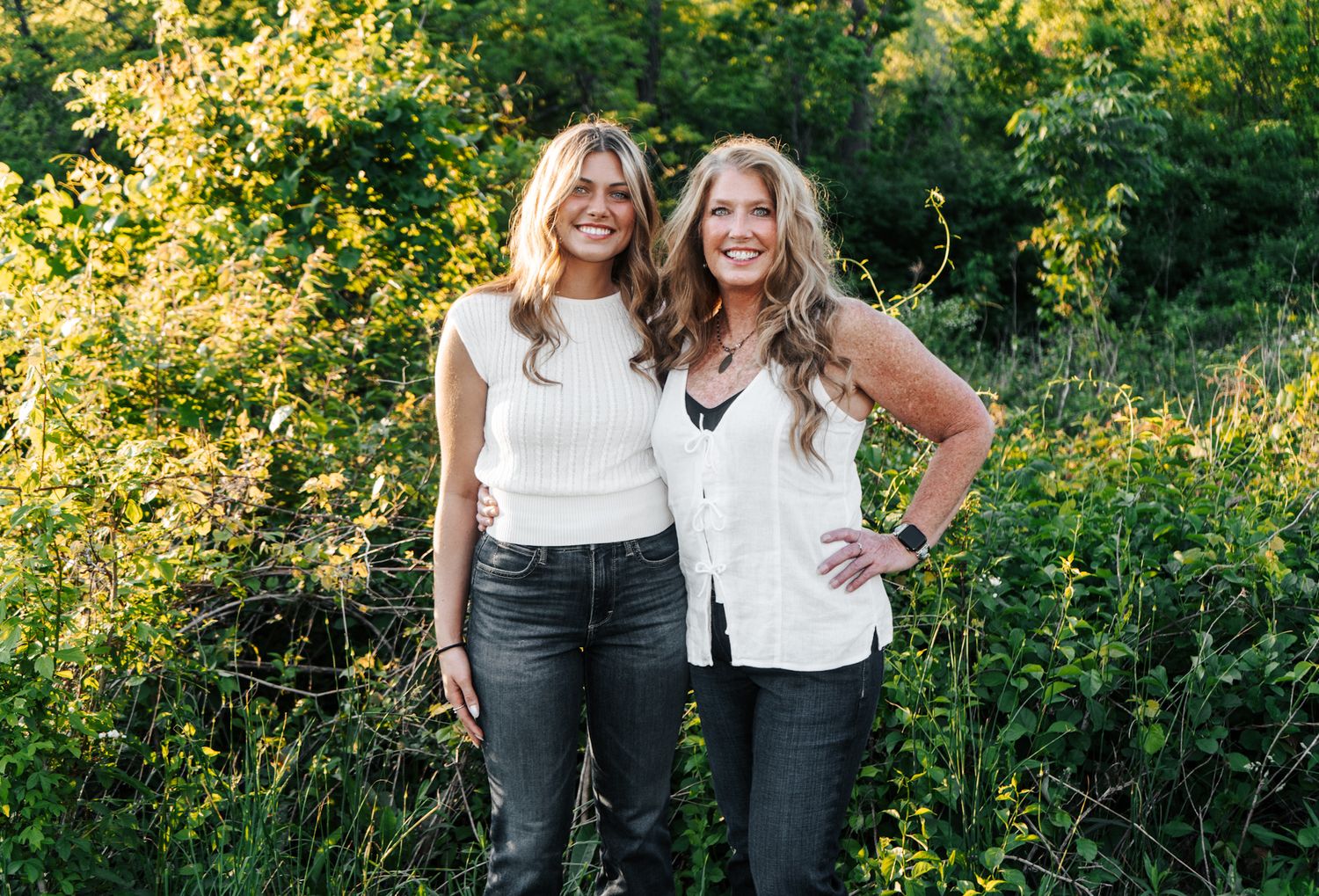 Two friends smiling together in casual outfits against lush summer foliage.