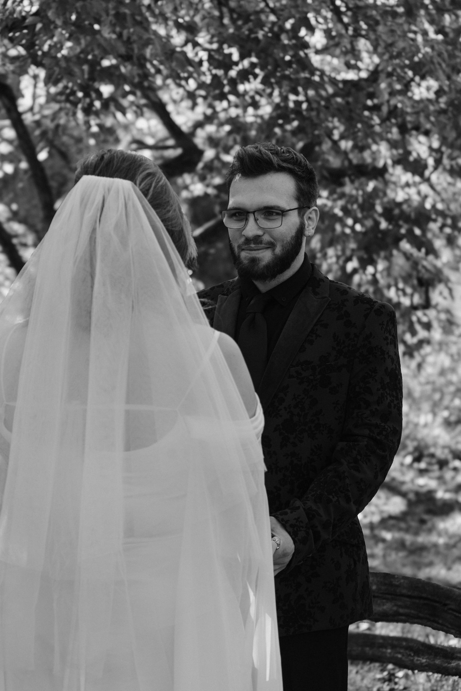 A black and white wedding photograph showing a couple during their outdoor ceremony beneath trees.