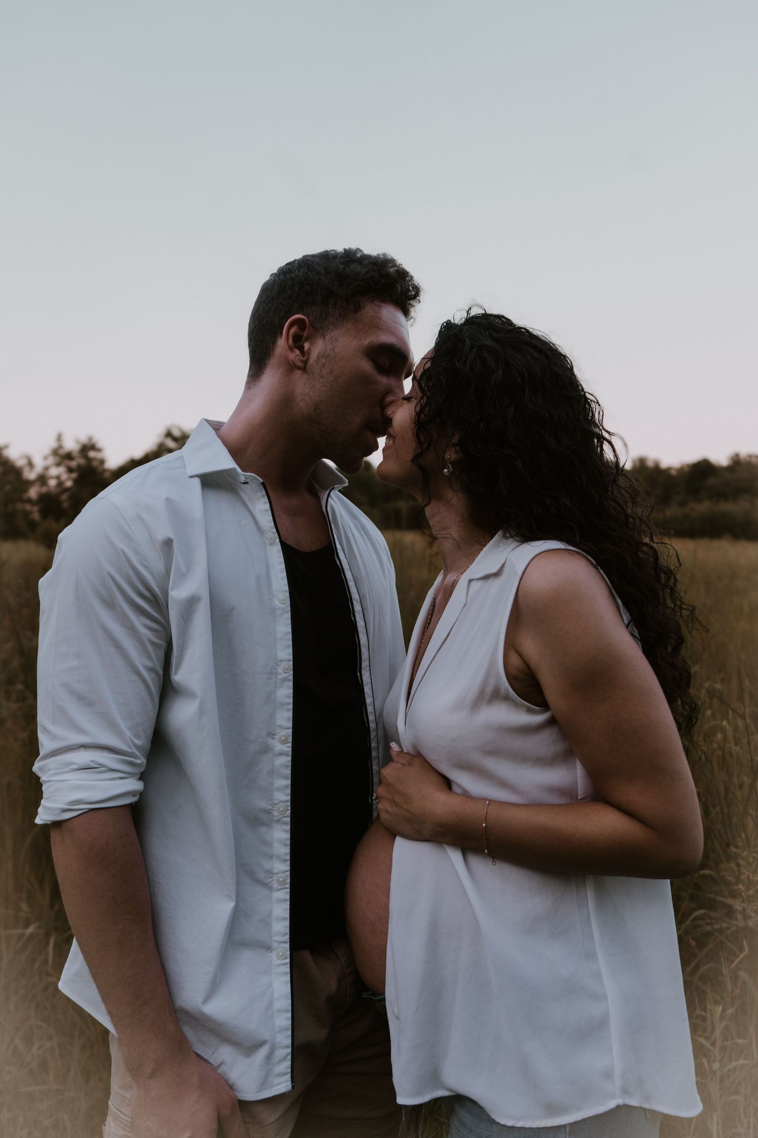 Romantic couple in white shirts sharing an intimate moment in a field at dusk.