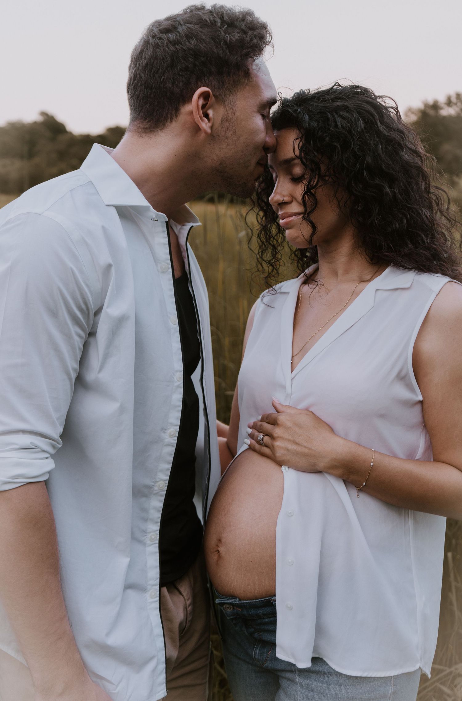Intimate couple standing in field with pregnant belly showing through white shirt and jeans at sunset.