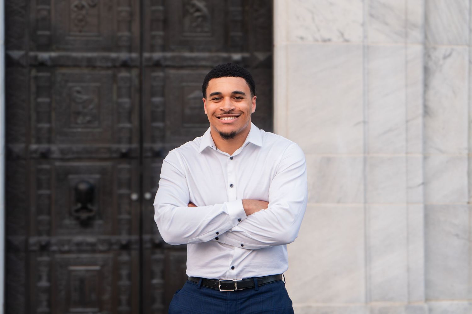Professional headshot against ornate door and white wall showing confident pose with arms crossed and wearing button-down shirt.