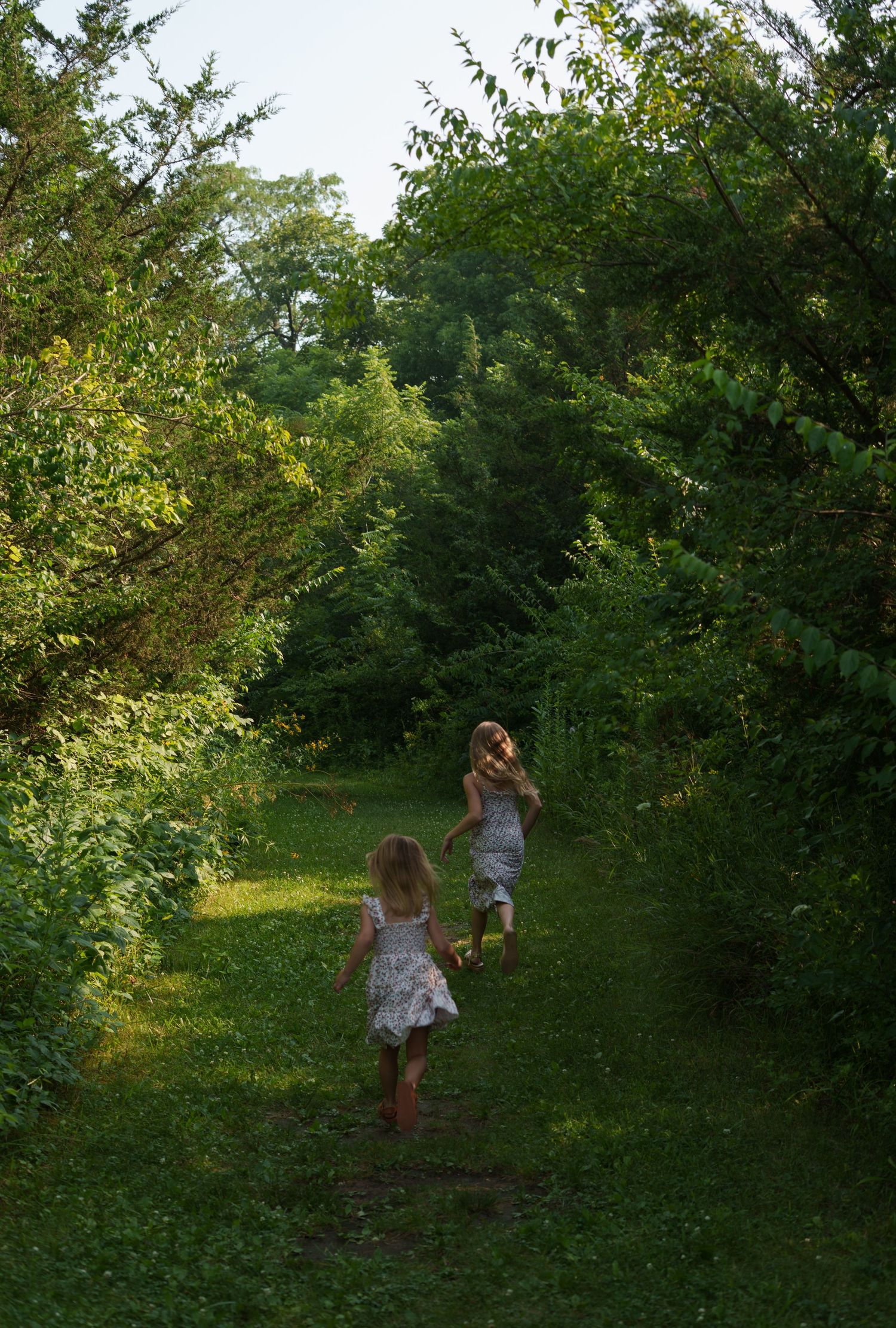 Two young girls in white dresses run barefoot along a lush green garden path surrounded by trees.