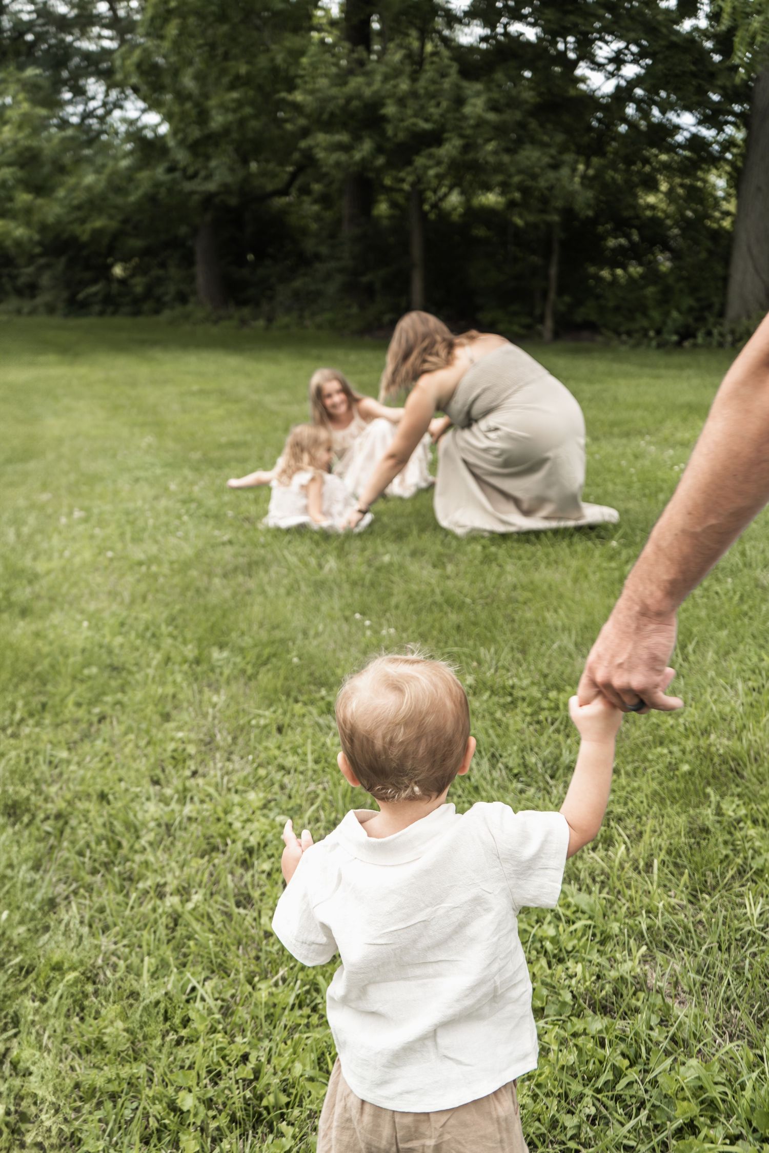 A small child holds an adult's hand while standing on grass as others sit in the background on a summer day.