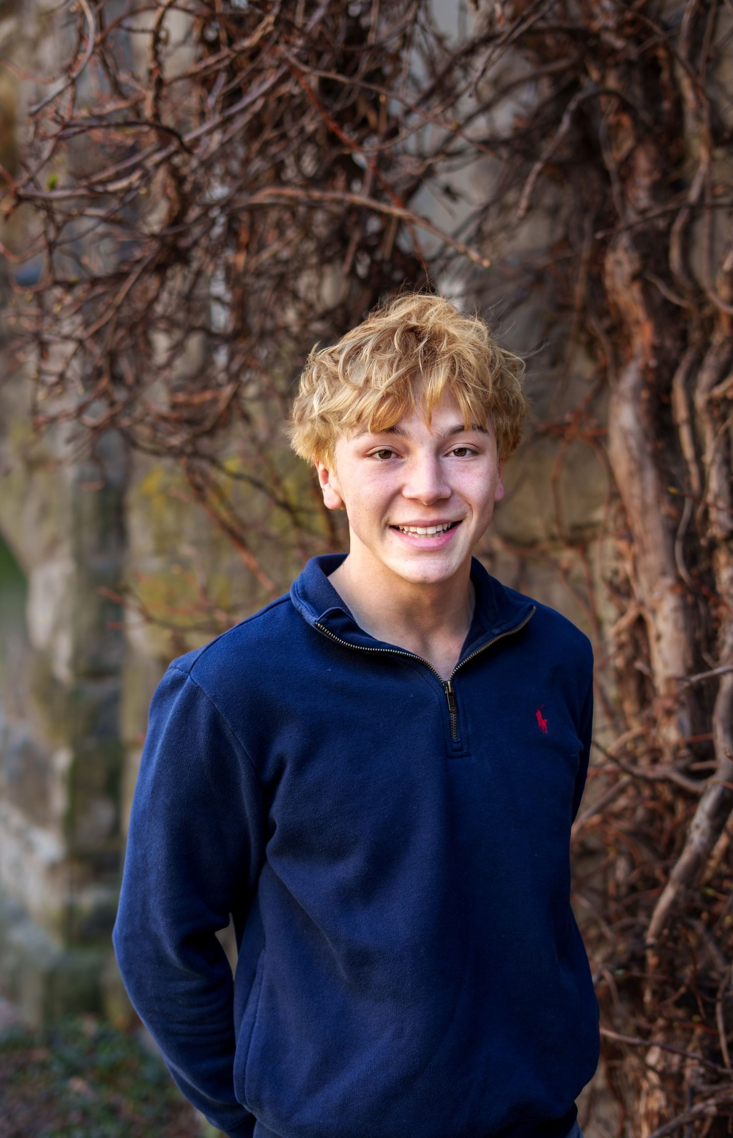 Smiling teenage boy with curly blonde hair wearing a navy Polo Ralph Lauren fleece jacket outdoors.