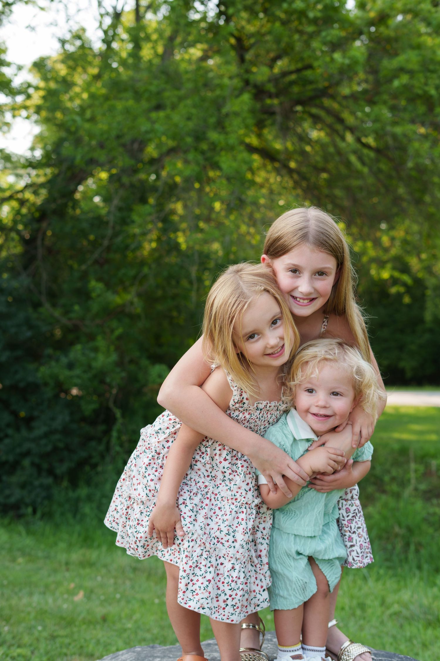 Three young sisters hugging and smiling outdoors in a lush green park, wearing summer dresses.