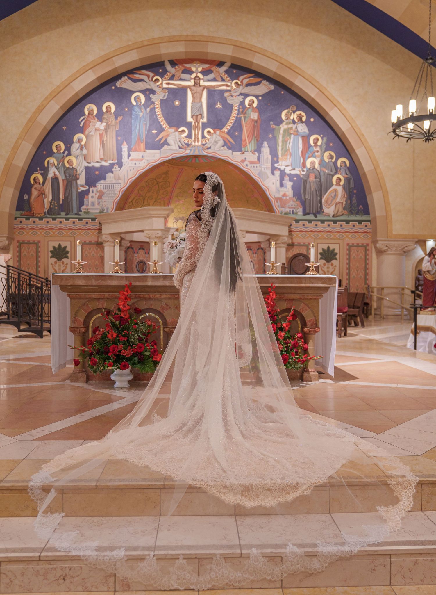 Bride in a long white gown and cathedral veil stands before ornate mosaic altar in a beautiful Catholic church.