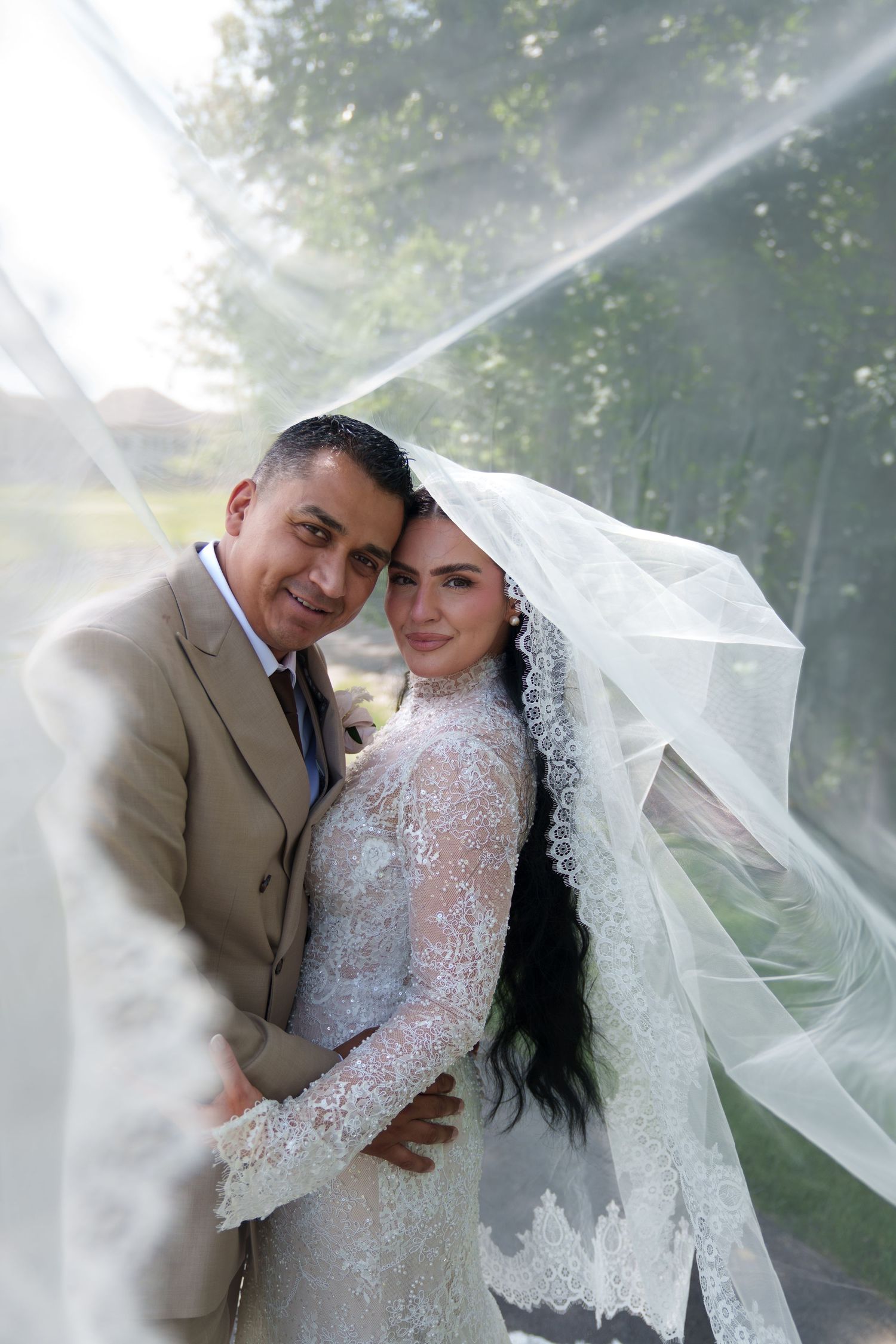 A bride in lace gown and veil embraces her groom in a tan suit outdoors on their wedding day.