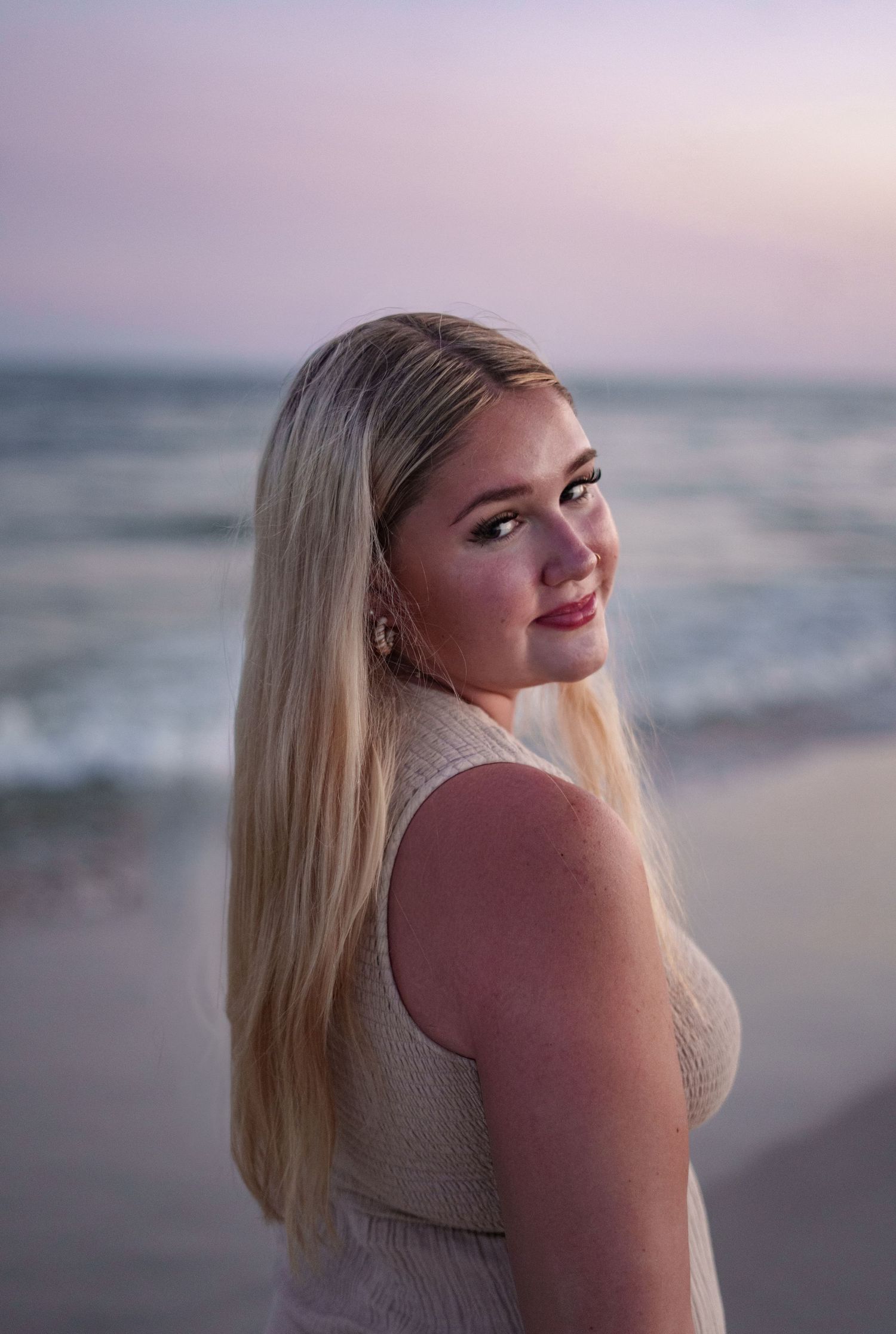 Young blonde woman smiling over shoulder at beach during sunset, wearing white dress with ocean waves in background.
