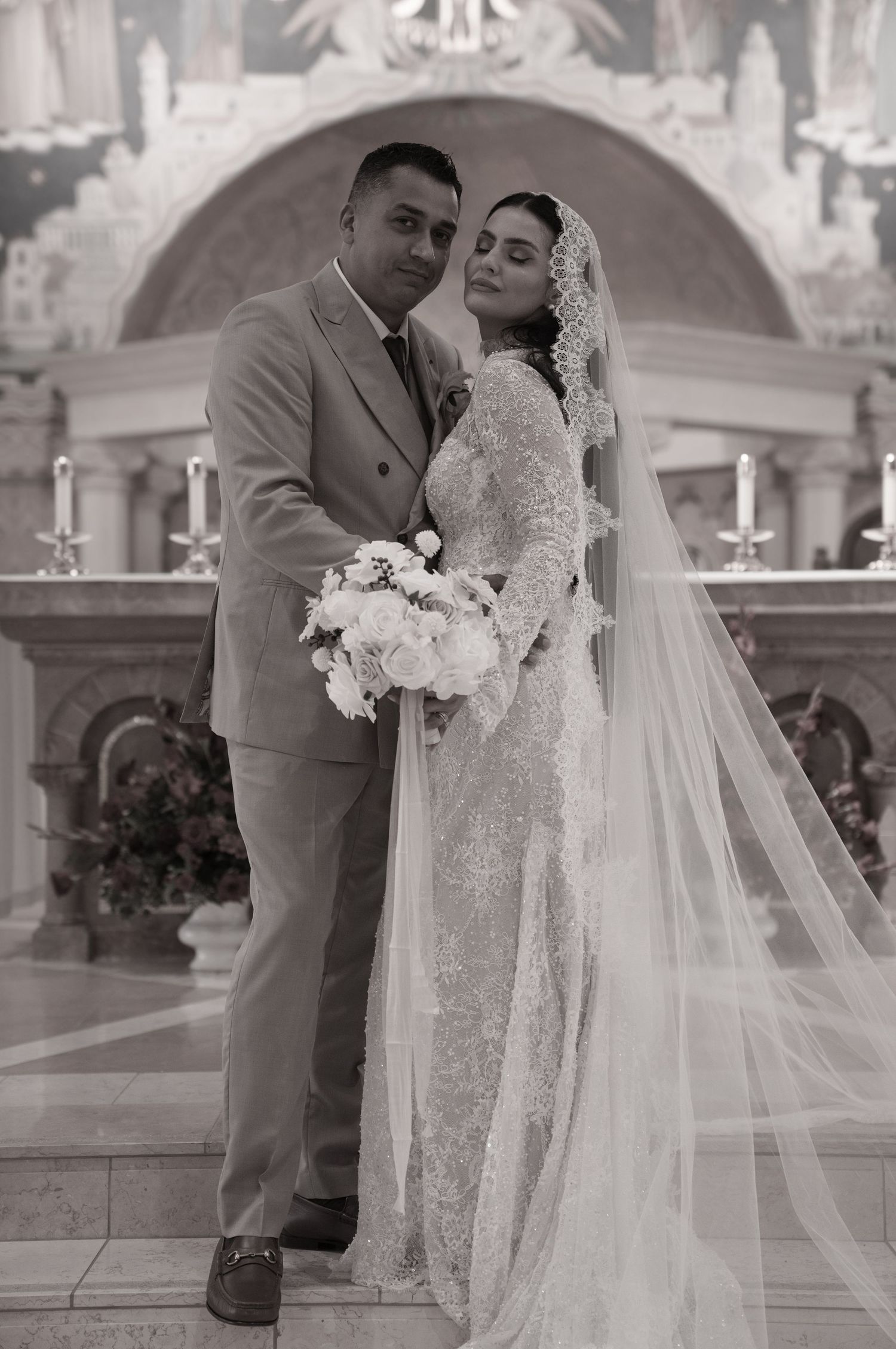 Black and white wedding photo of bride in lace gown with veil and groom in suit posing in elegant church.