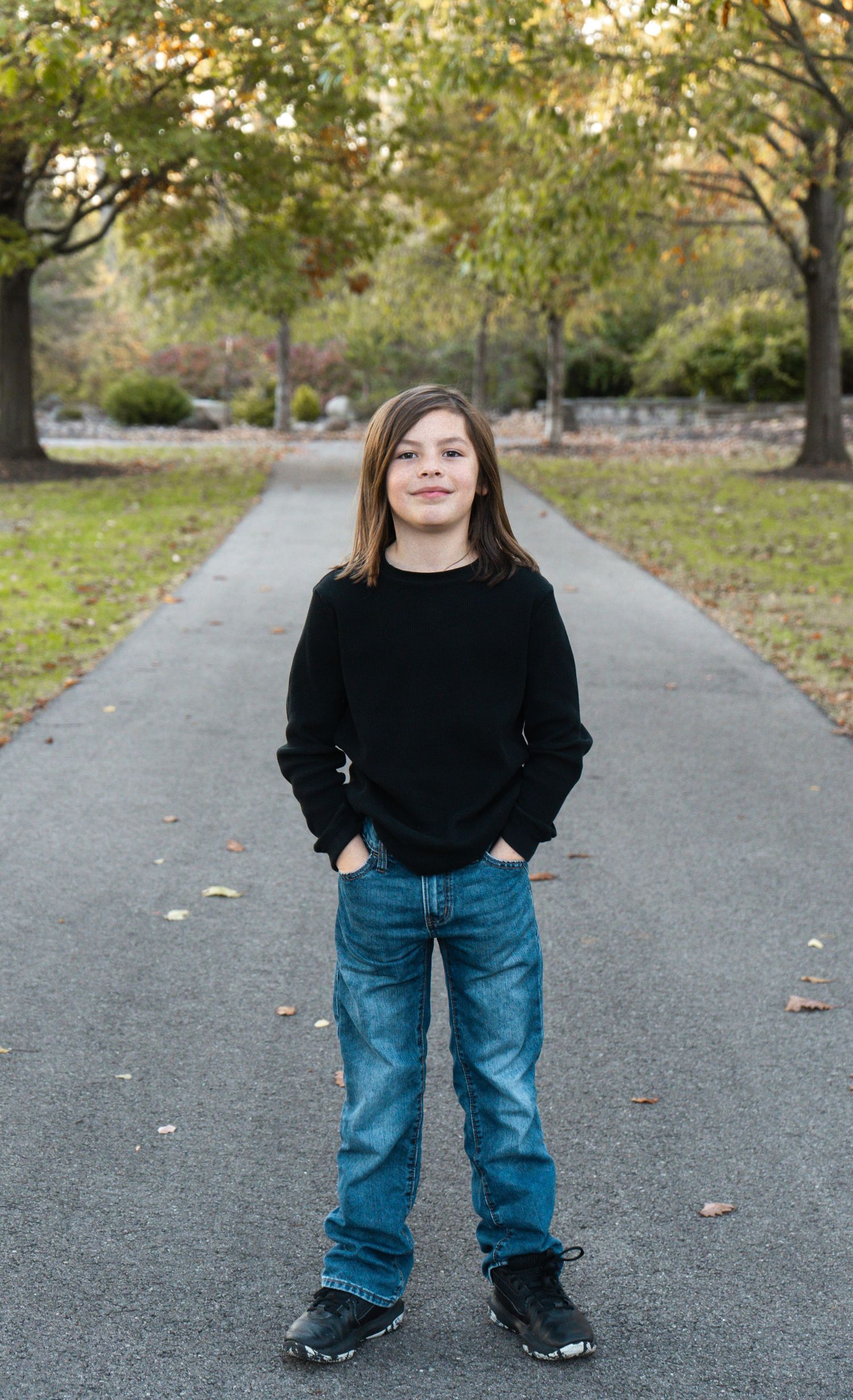 A person wearing a black sweater and jeans stands on a paved path surrounded by autumn trees and fallen leaves.