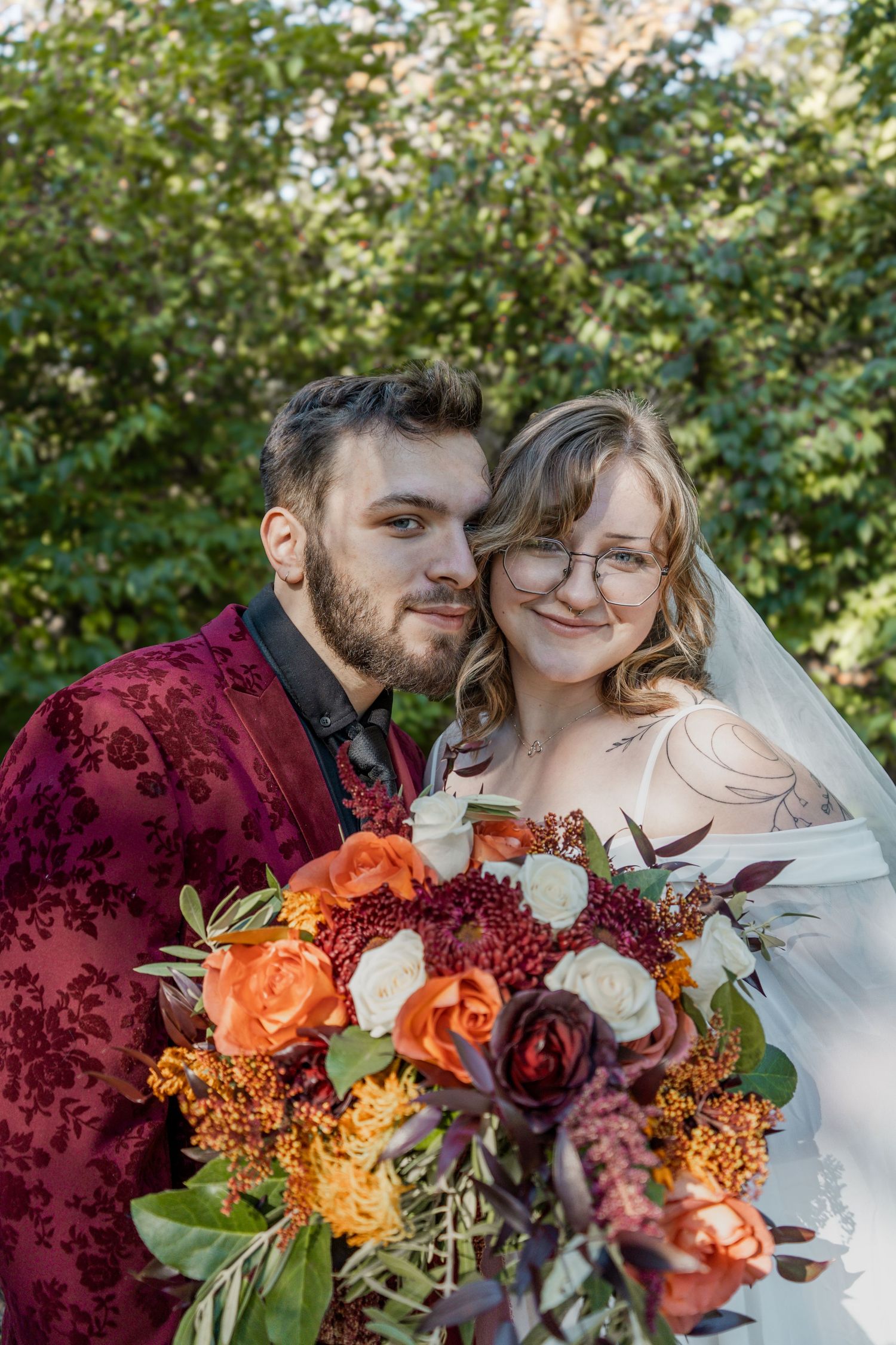 A bride and groom pose together outdoors, holding a vibrant autumn bouquet with orange and burgundy roses.