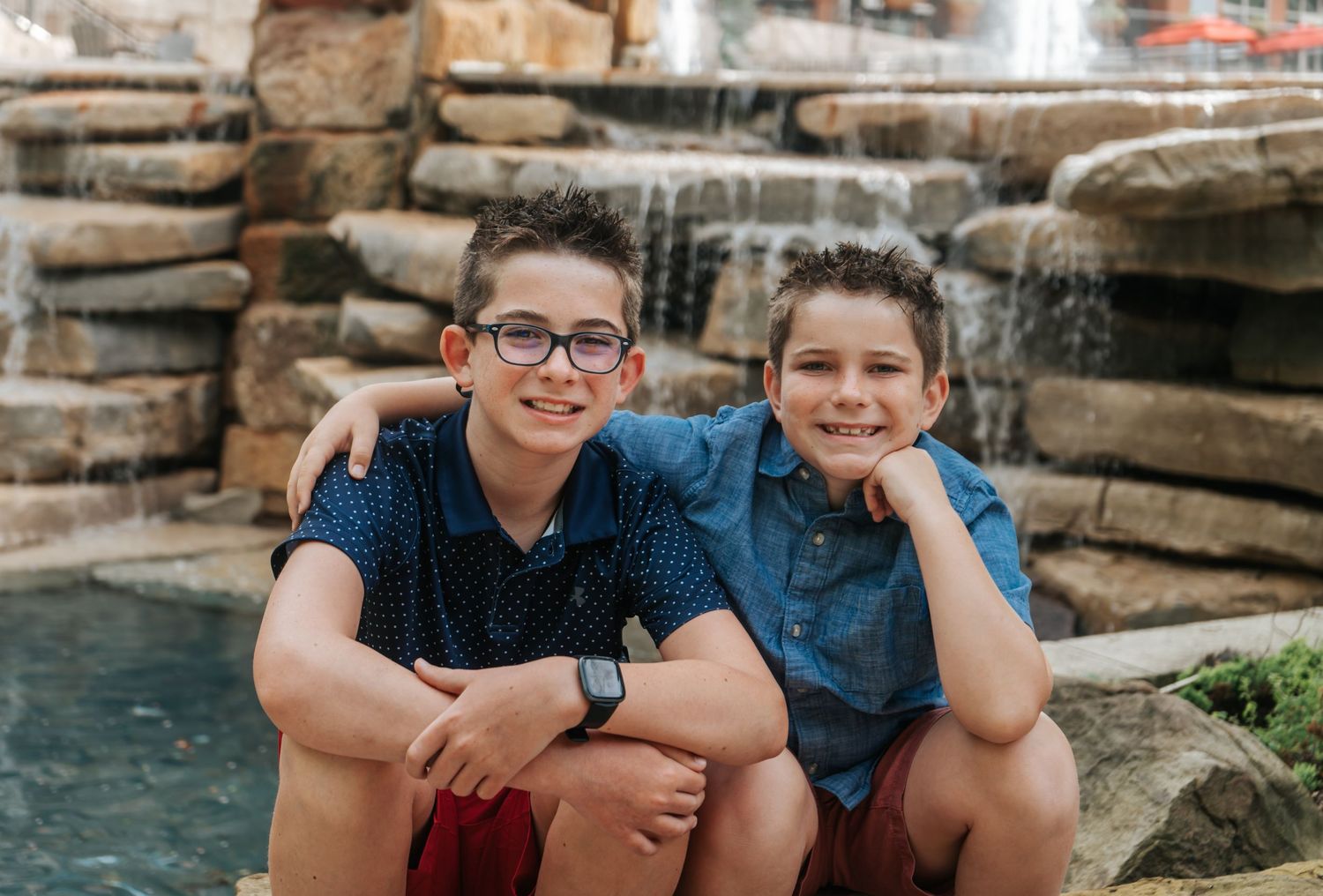 Two young people in casual blue shirts sit together by a decorative rock waterfall feature in an outdoor setting.