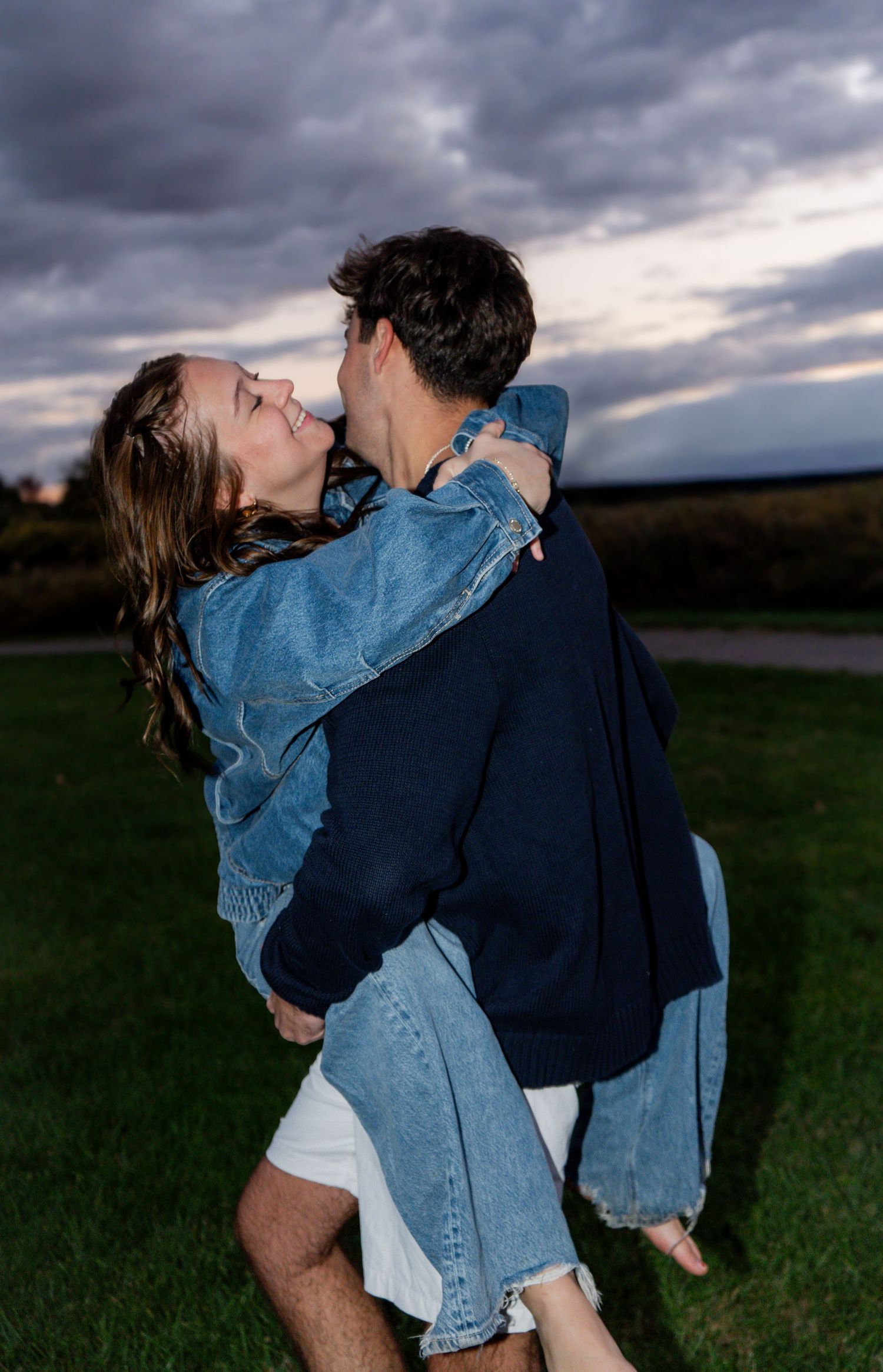 A romantic couple embraces in a field at dusk with dramatic storm clouds overhead.