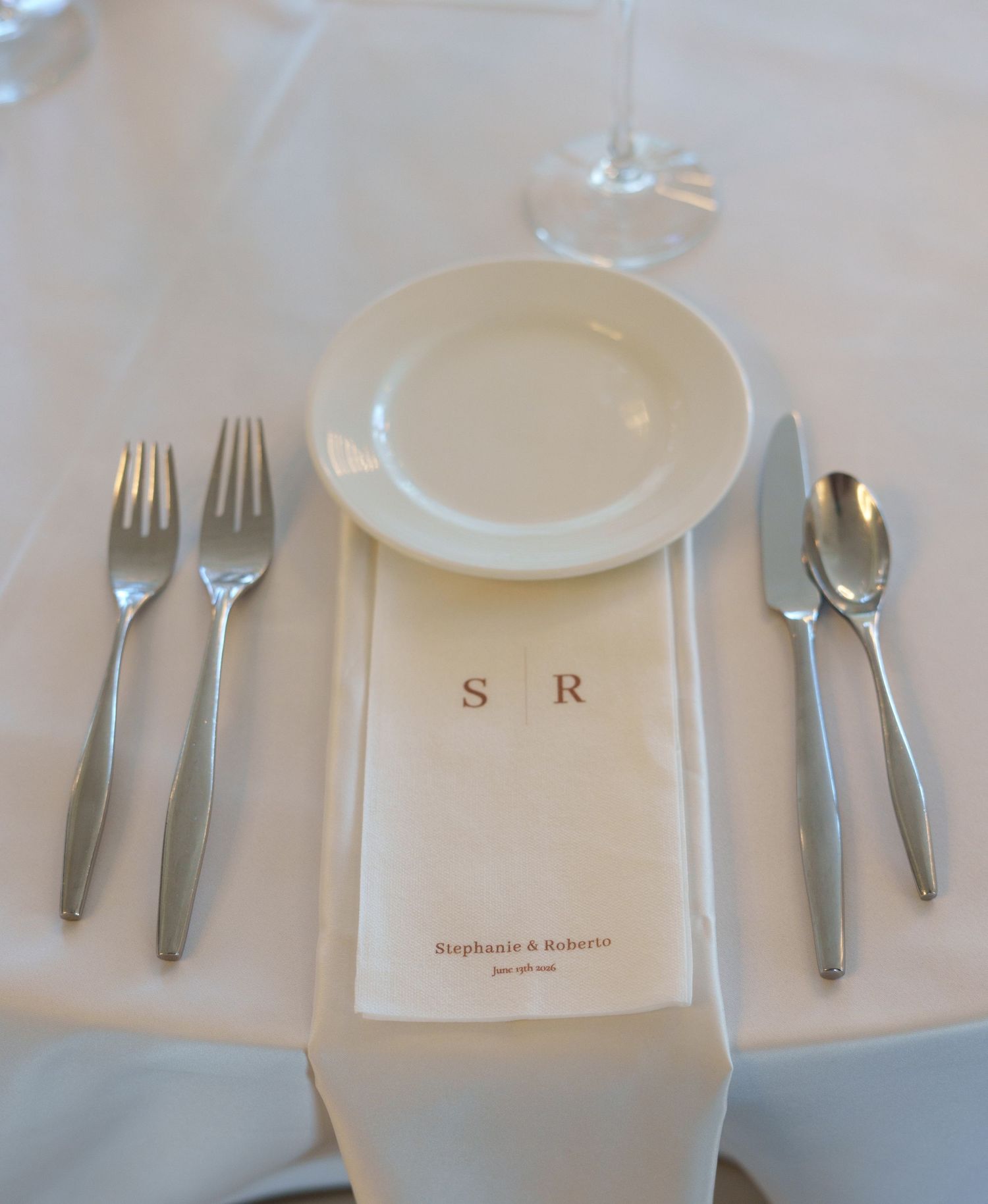 Elegant wedding place setting with monogrammed napkin for Stephanie & Roberto, two forks, knife, spoon, plate, and wine glass.