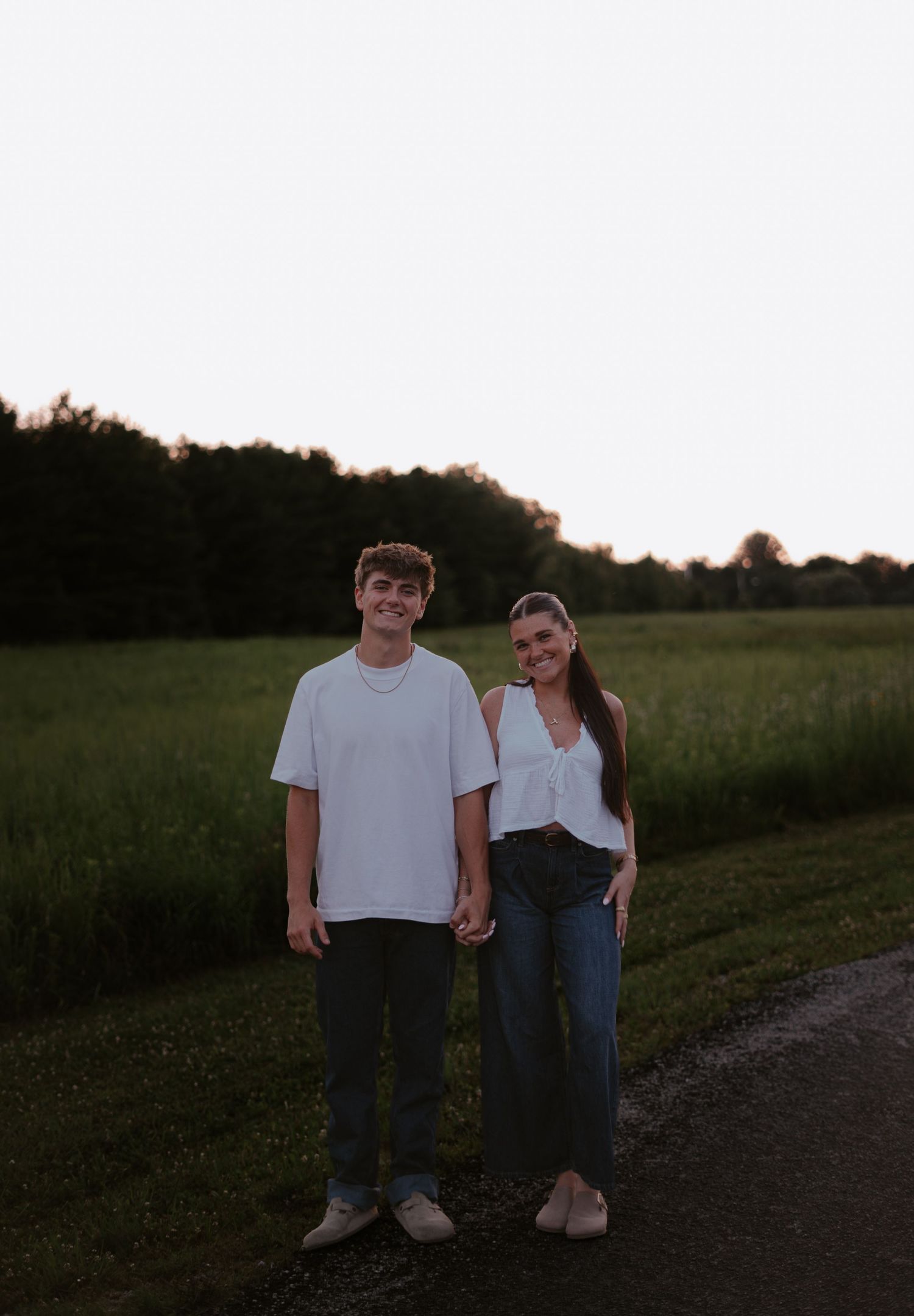 A young couple holds hands and smiles while standing on a rural path at sunset with green fields and trees behind them.