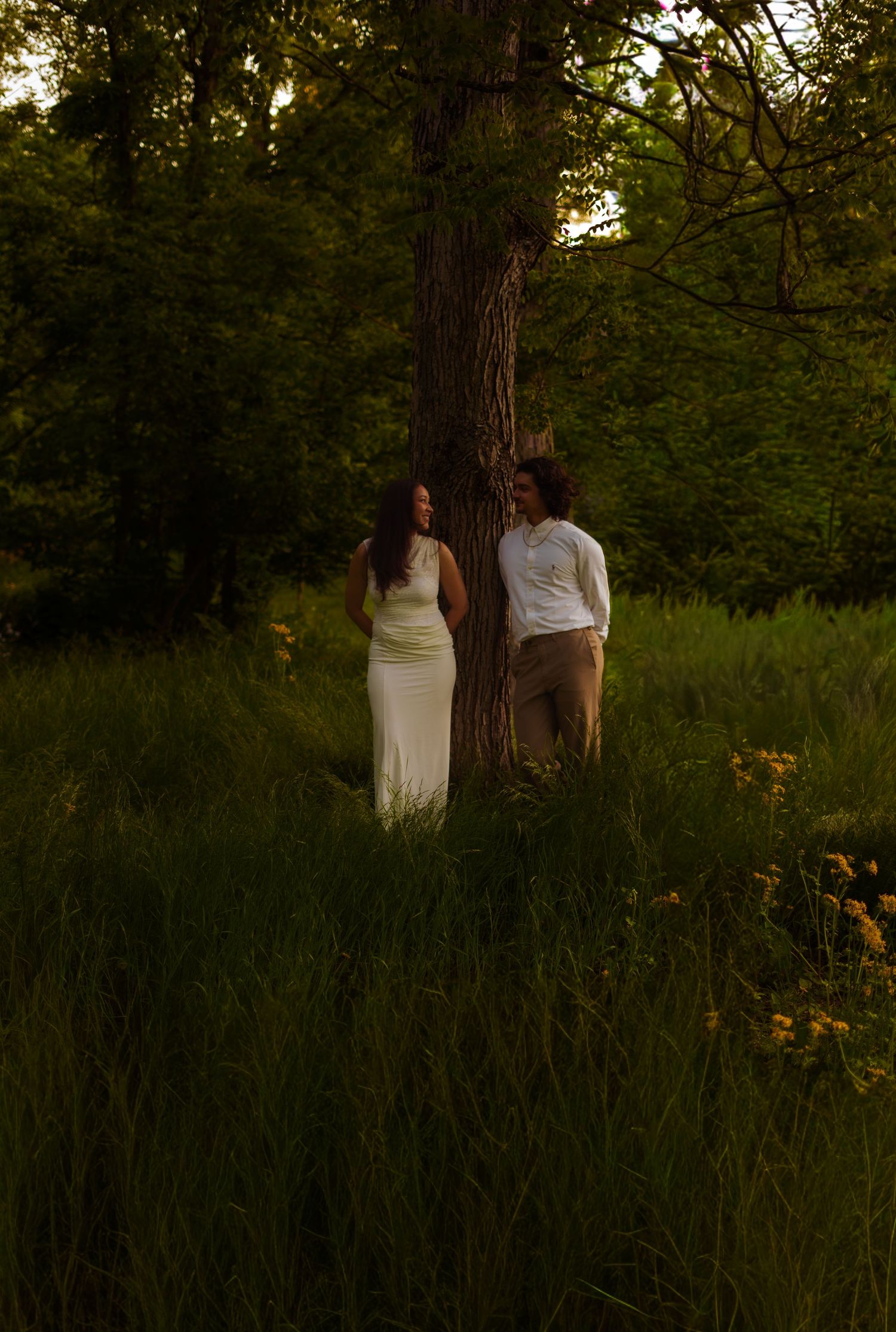 A couple stands on opposite sides of a tree in a lush green meadow, the woman in white dress, man in casual attire.