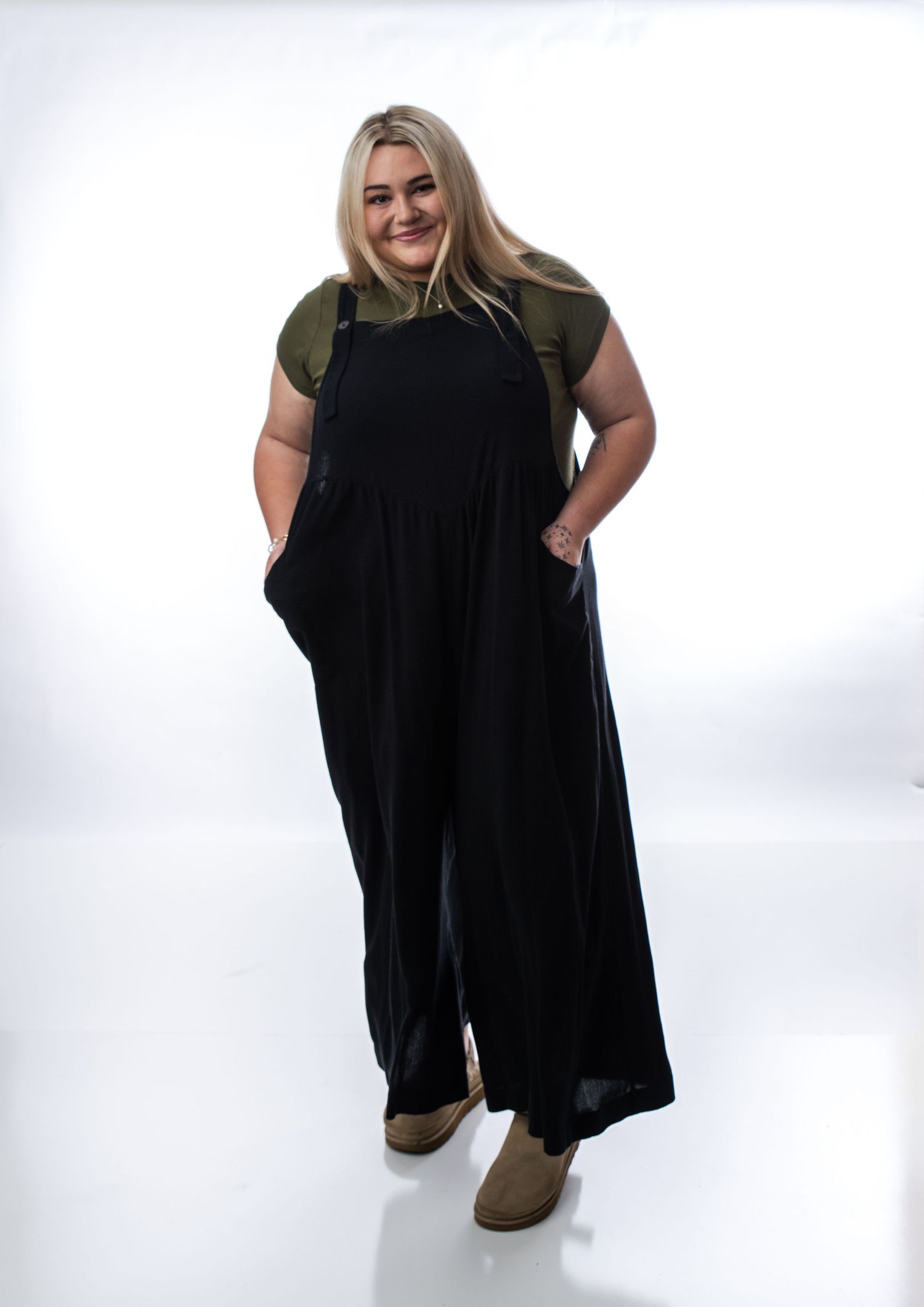 Plus size woman wearing black wide-leg overalls over olive green tee with tan ankle boots, posed on white background.