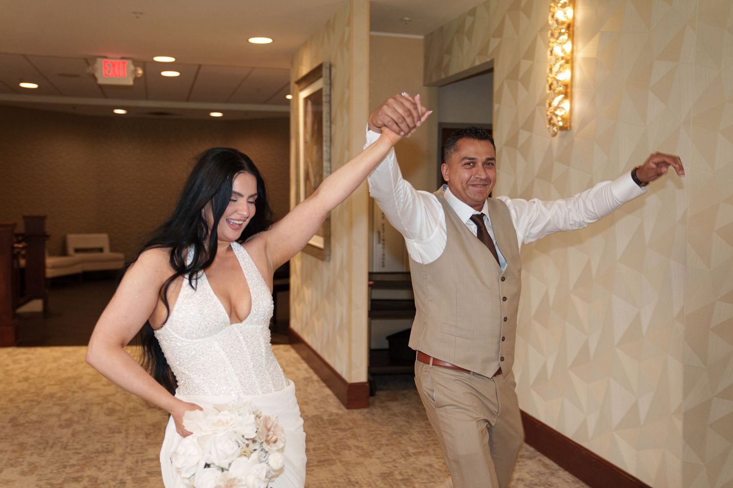 Bride and groom dancing joyfully in wedding attire, bride holding bouquet, in elegant hotel ballroom.