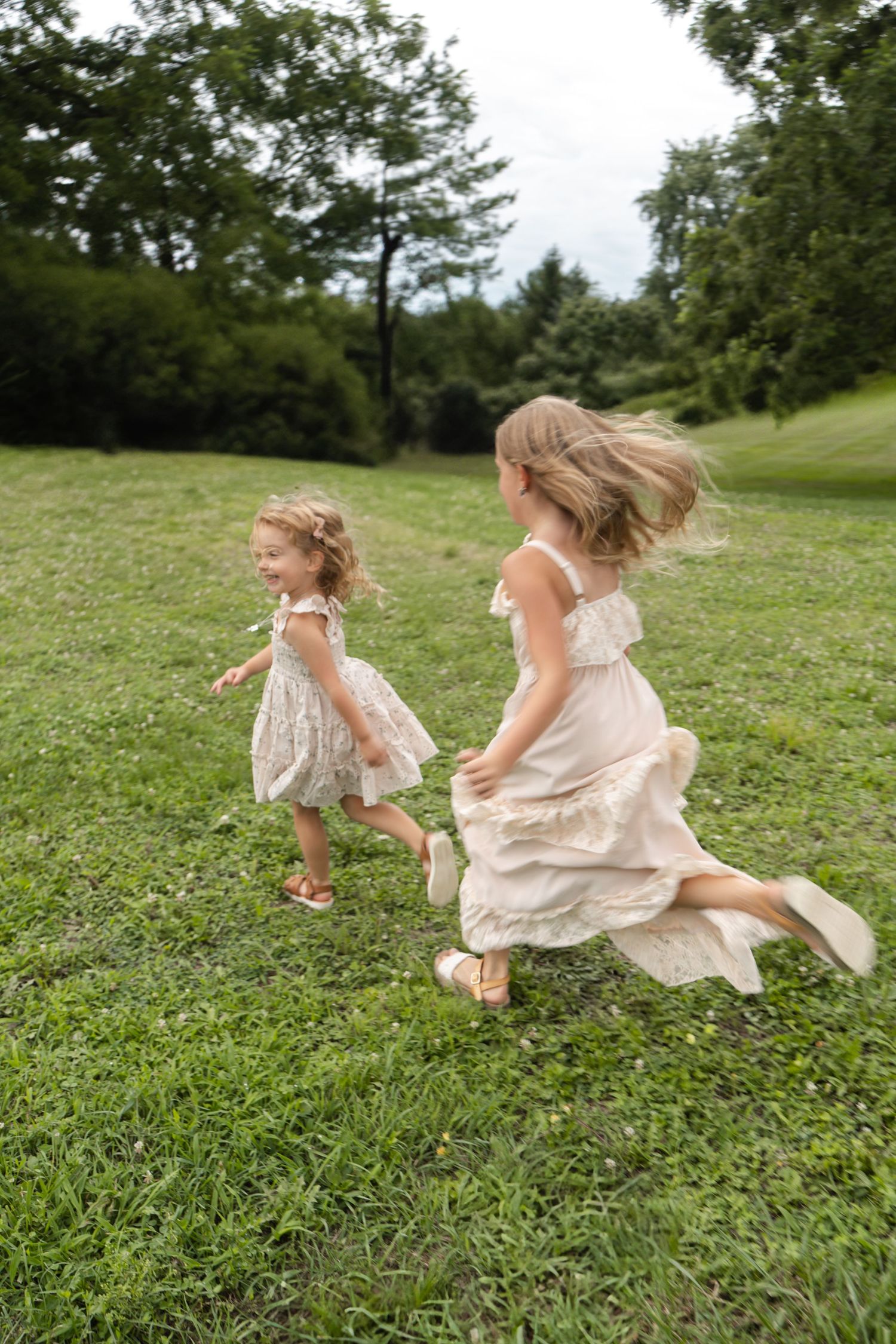 Two figures in flowing white dresses run and play together on a grassy hillside on a summer day.
