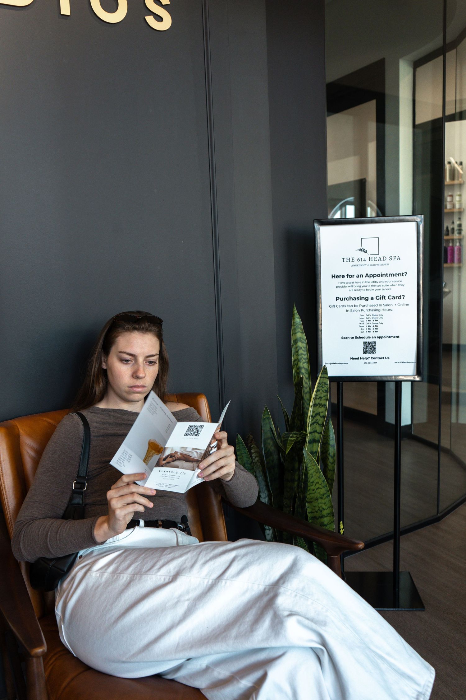 A person in white loungewear reclines on a brown leather chair while reading a document in an office setting.