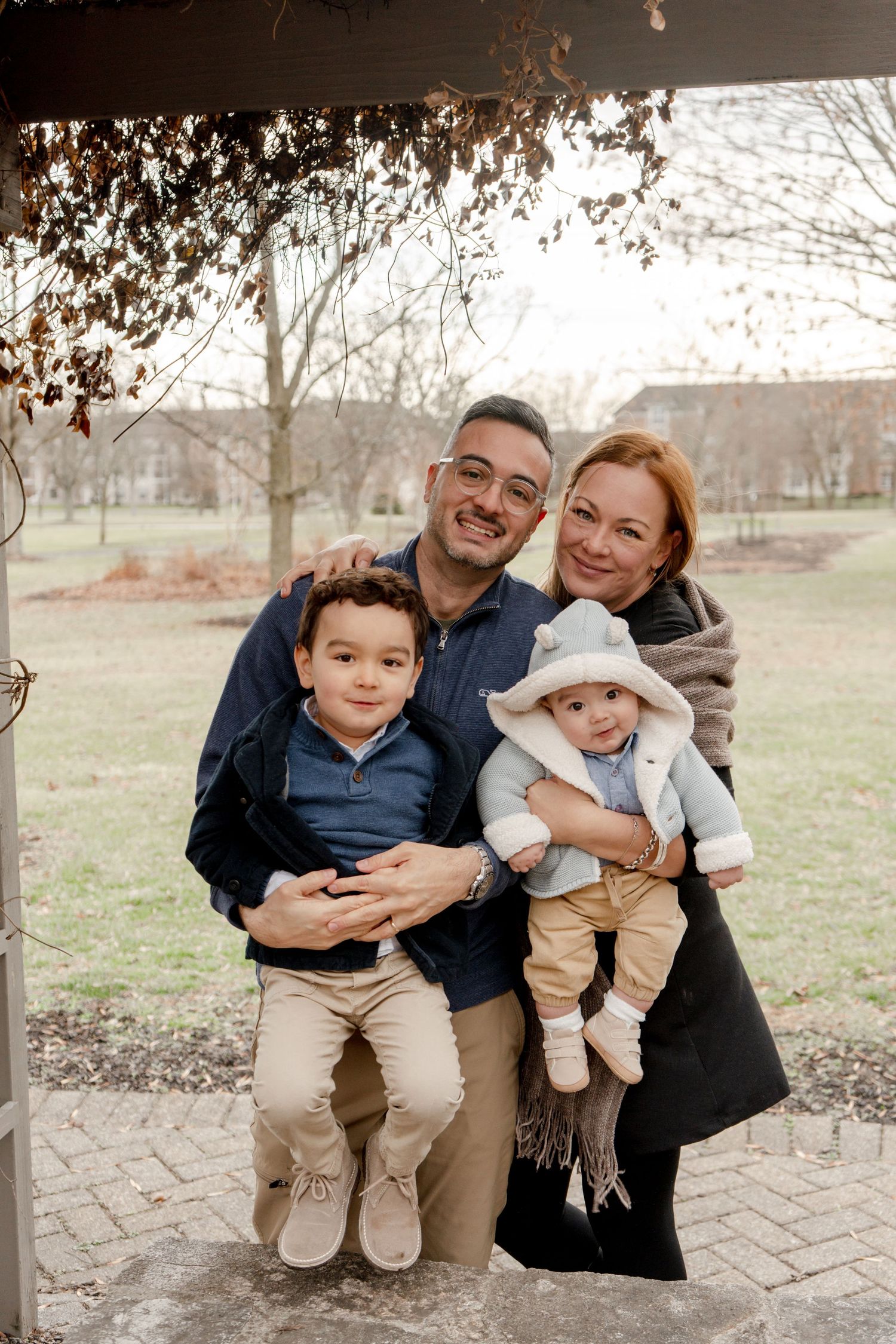Family of four posing outdoors in autumn, parents holding two young boys, smiling warmly in a park setting.