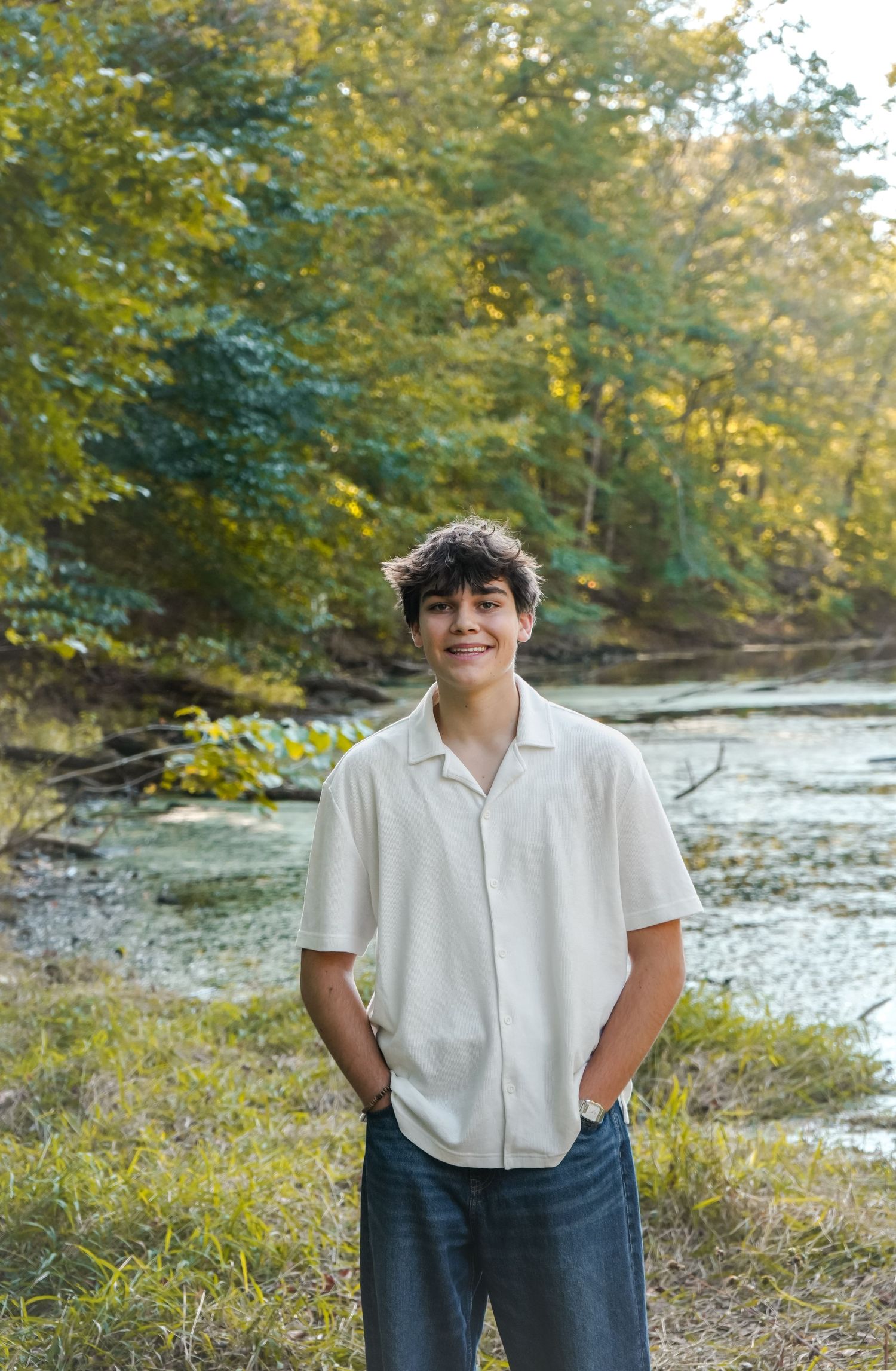 Smiling teenage boy in white shirt and jeans standing outdoors near a creek surrounded by lush green trees.