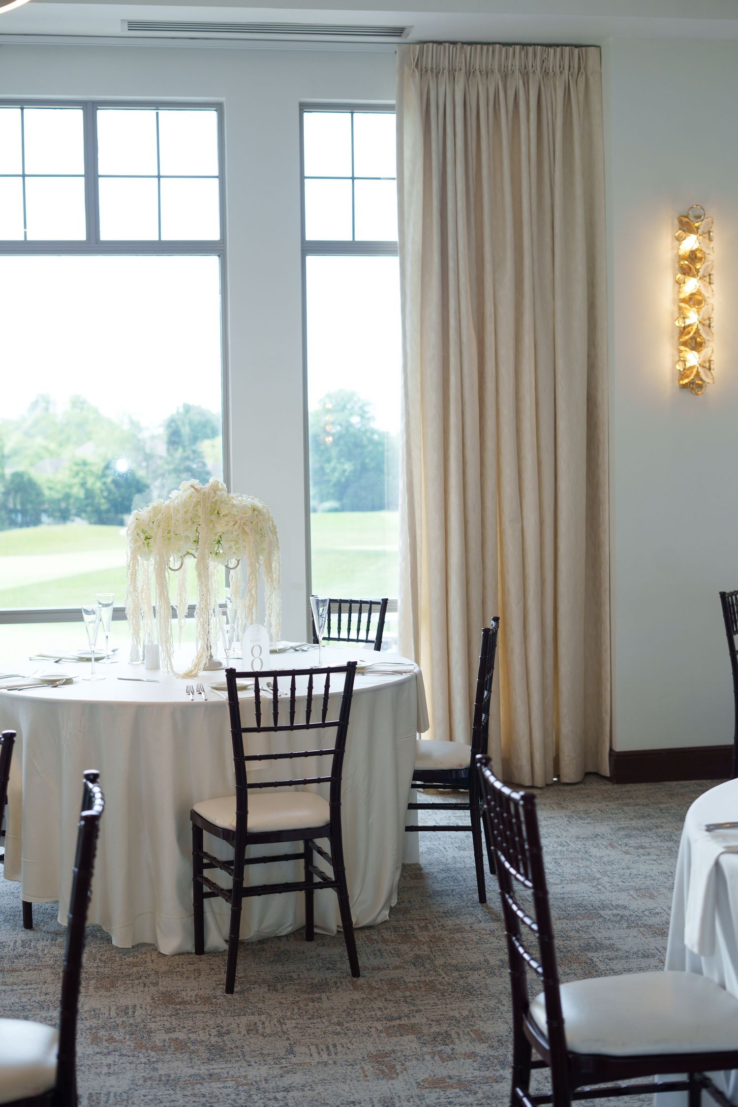 Elegant wedding reception table with white linen, floral centerpiece, chiavari chairs, and large windows overlooking a golf course.