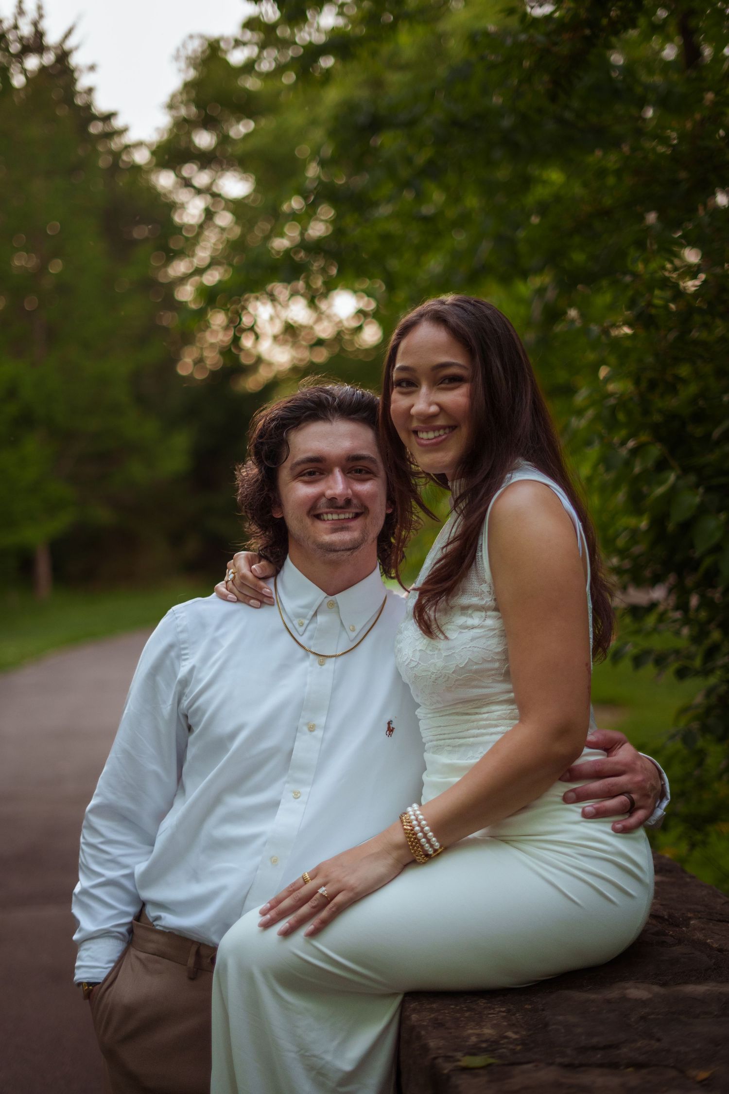 A smiling couple in white outfits poses together on a stone wall surrounded by lush green trees.