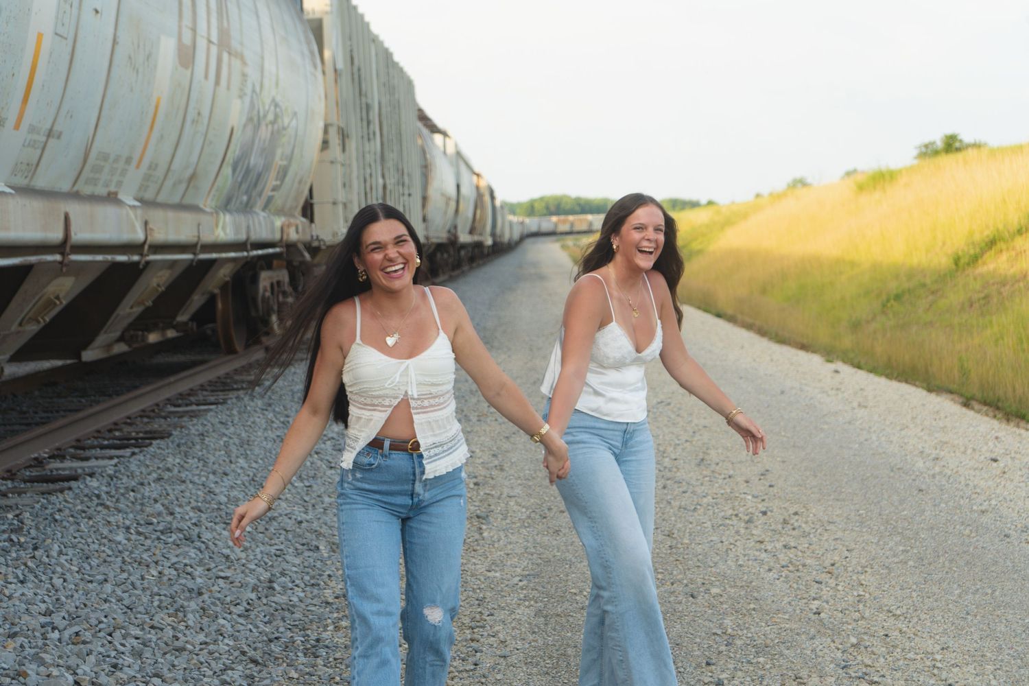 Two friends in white tops and jeans stroll and laugh together beside parked train cars on gravel path at sunset.