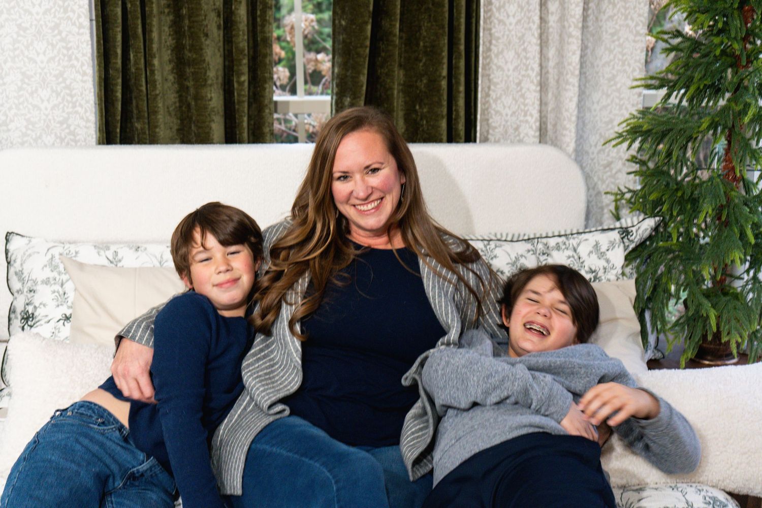 A family lounges together on a white couch, smiling and embracing while wearing casual blue and gray clothing.