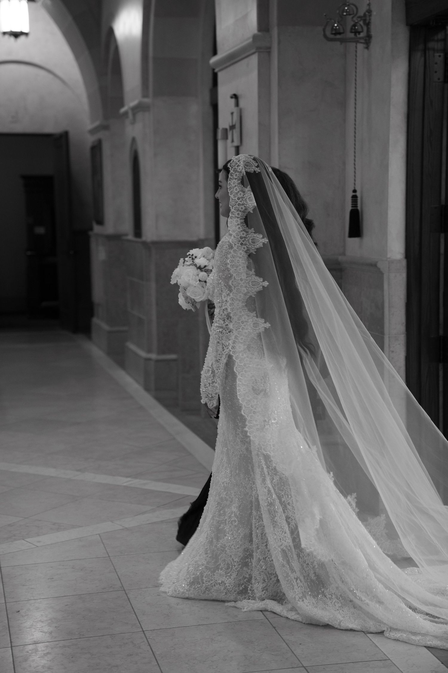 Black and white photo of a bride in lace gown with long veil holding bouquet walking through elegant church corridor.