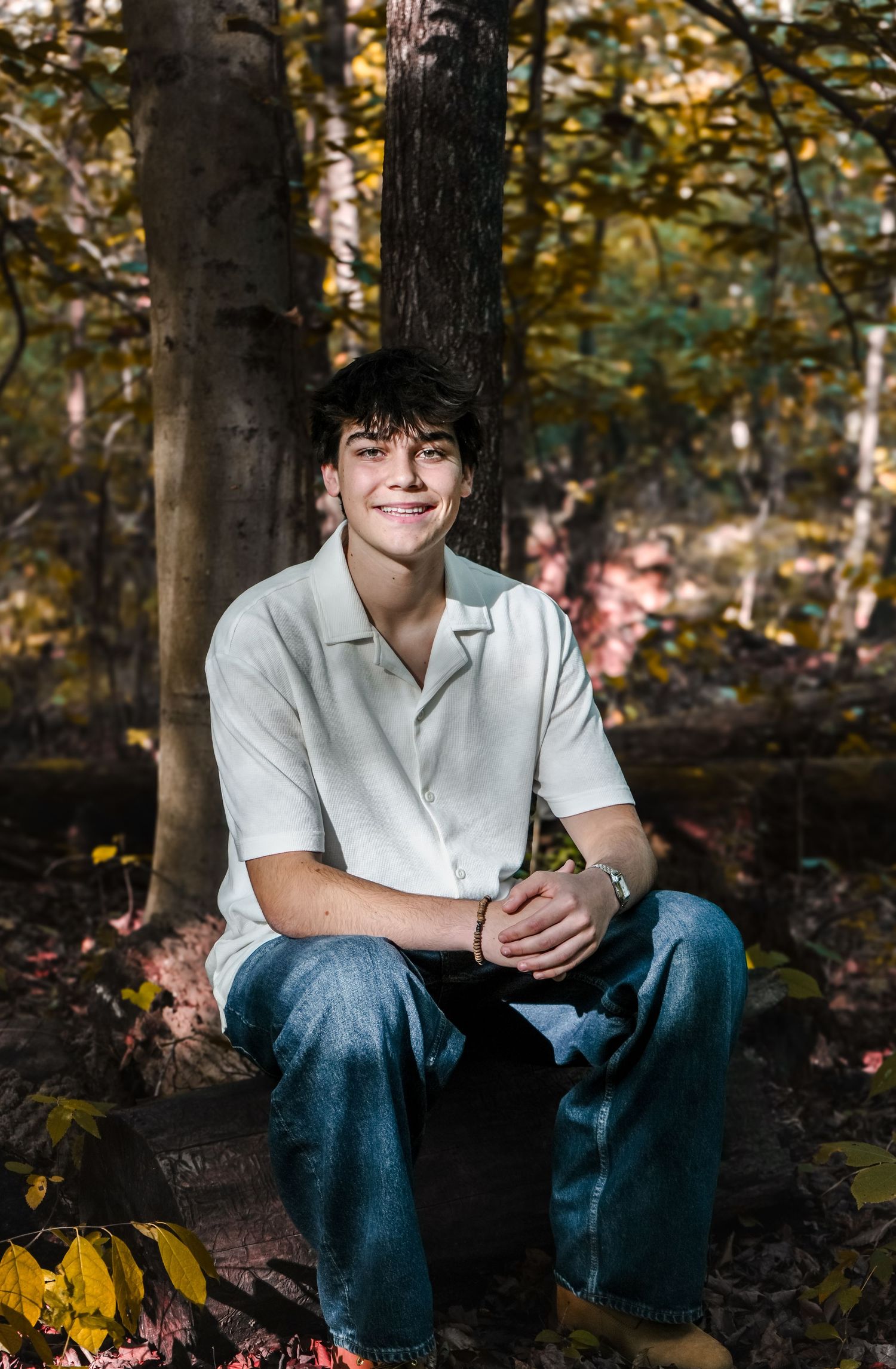 A smiling teenage boy sits on a log in an autumn forest, wearing a white shirt and jeans.