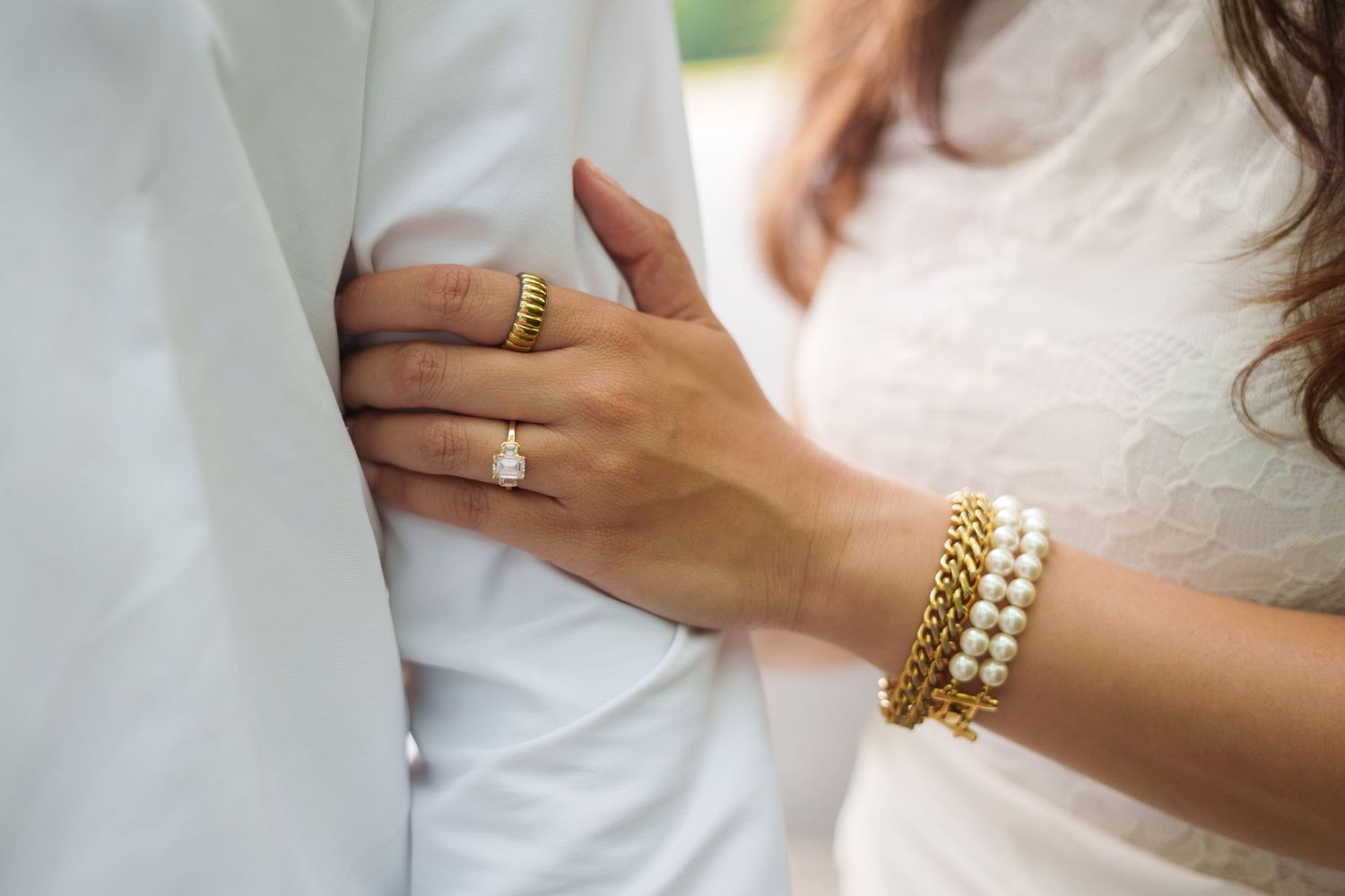 Close-up of bride's hand with diamond engagement ring and gold bracelets resting on groom's white suit jacket.