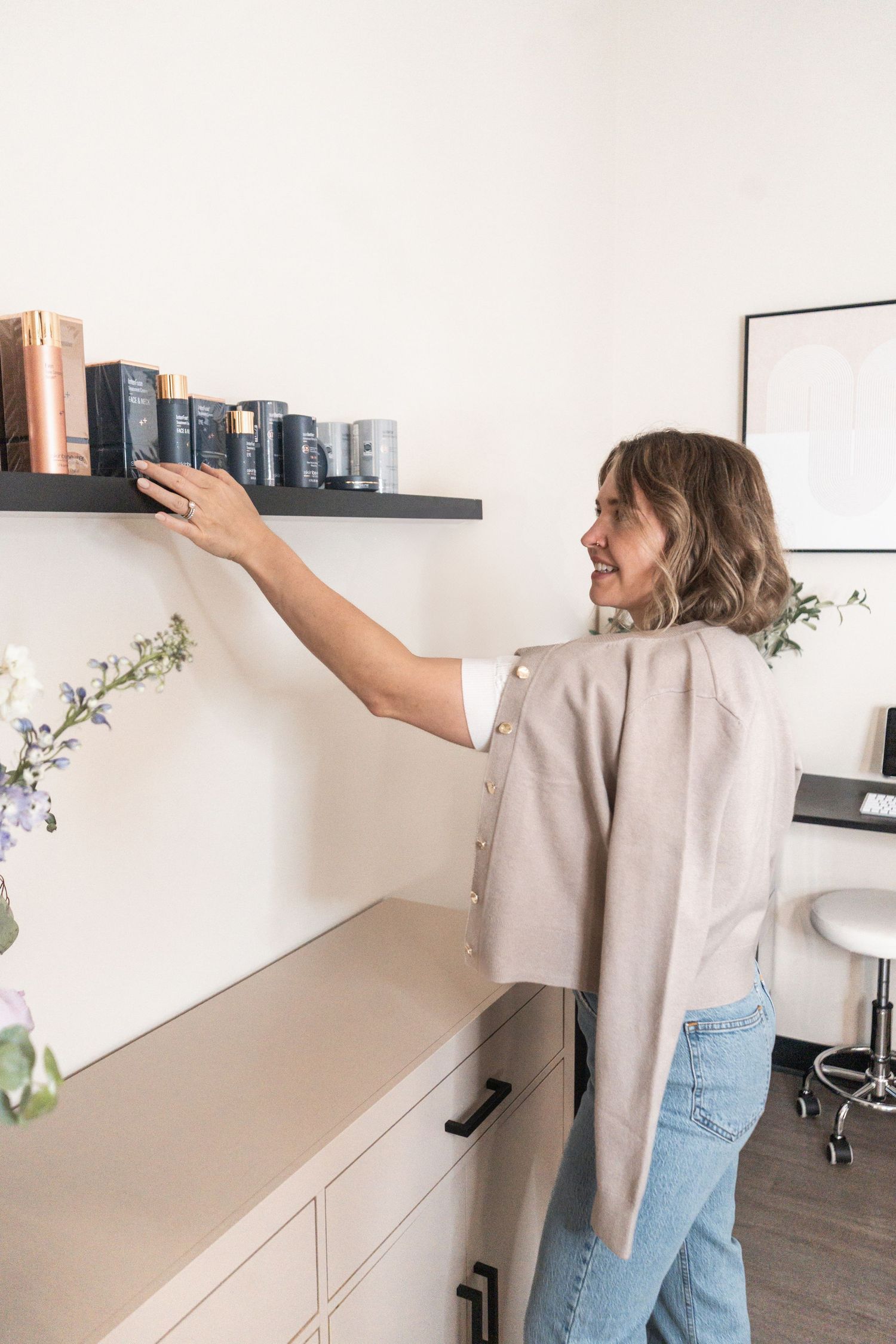 Someone in casual wear reaches for books on a floating shelf above white cabinets in a minimalist home office space.