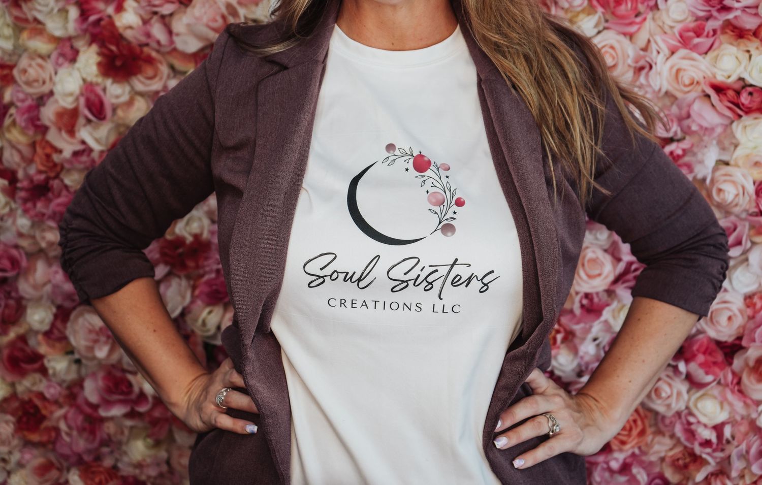 A person wearing a brown cardigan and white t-shirt with Sand Sisters logo against a floral backdrop.