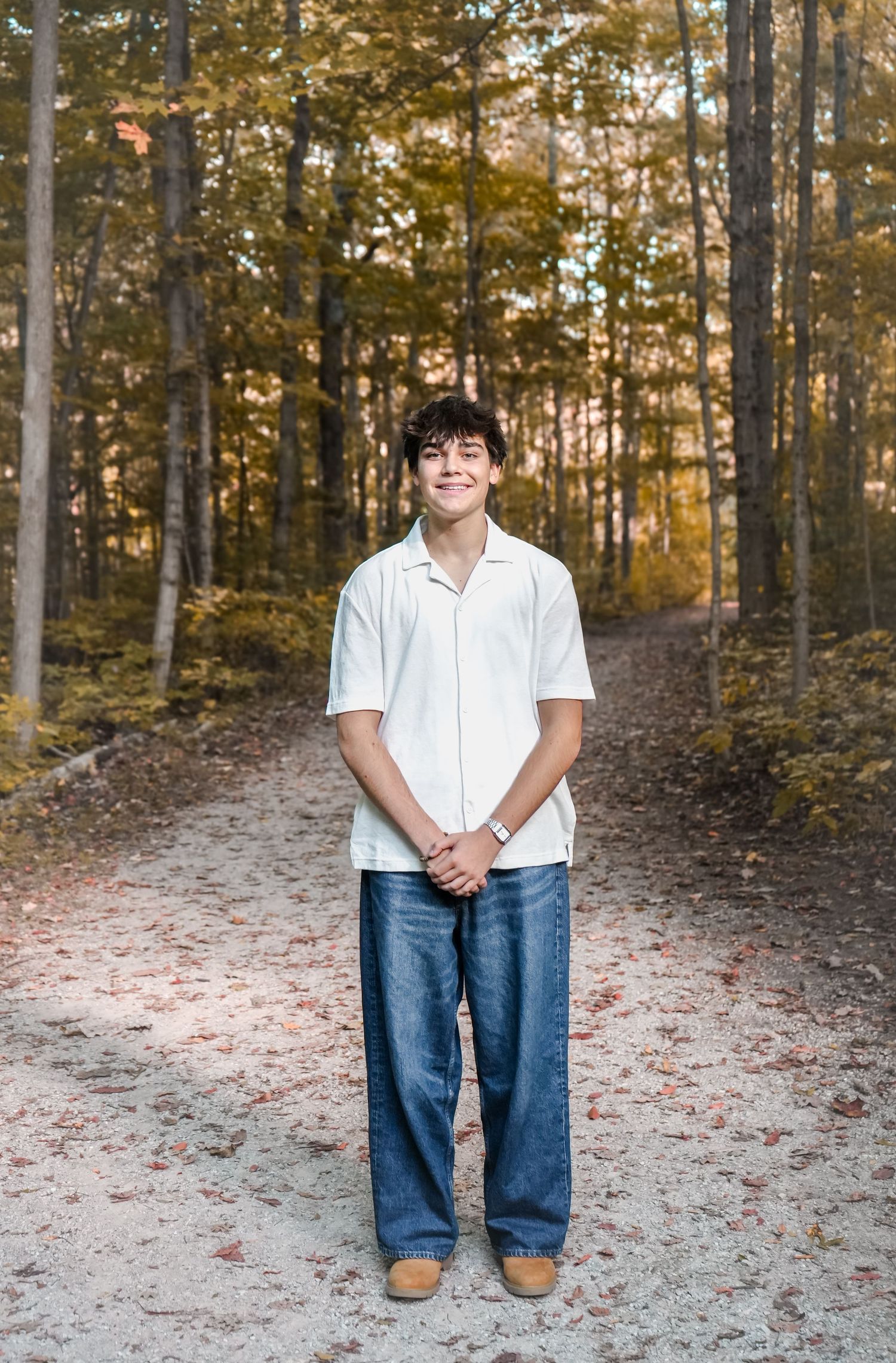 A person in a white shirt and jeans stands on a forest trail during autumn with golden foliage in the background.
