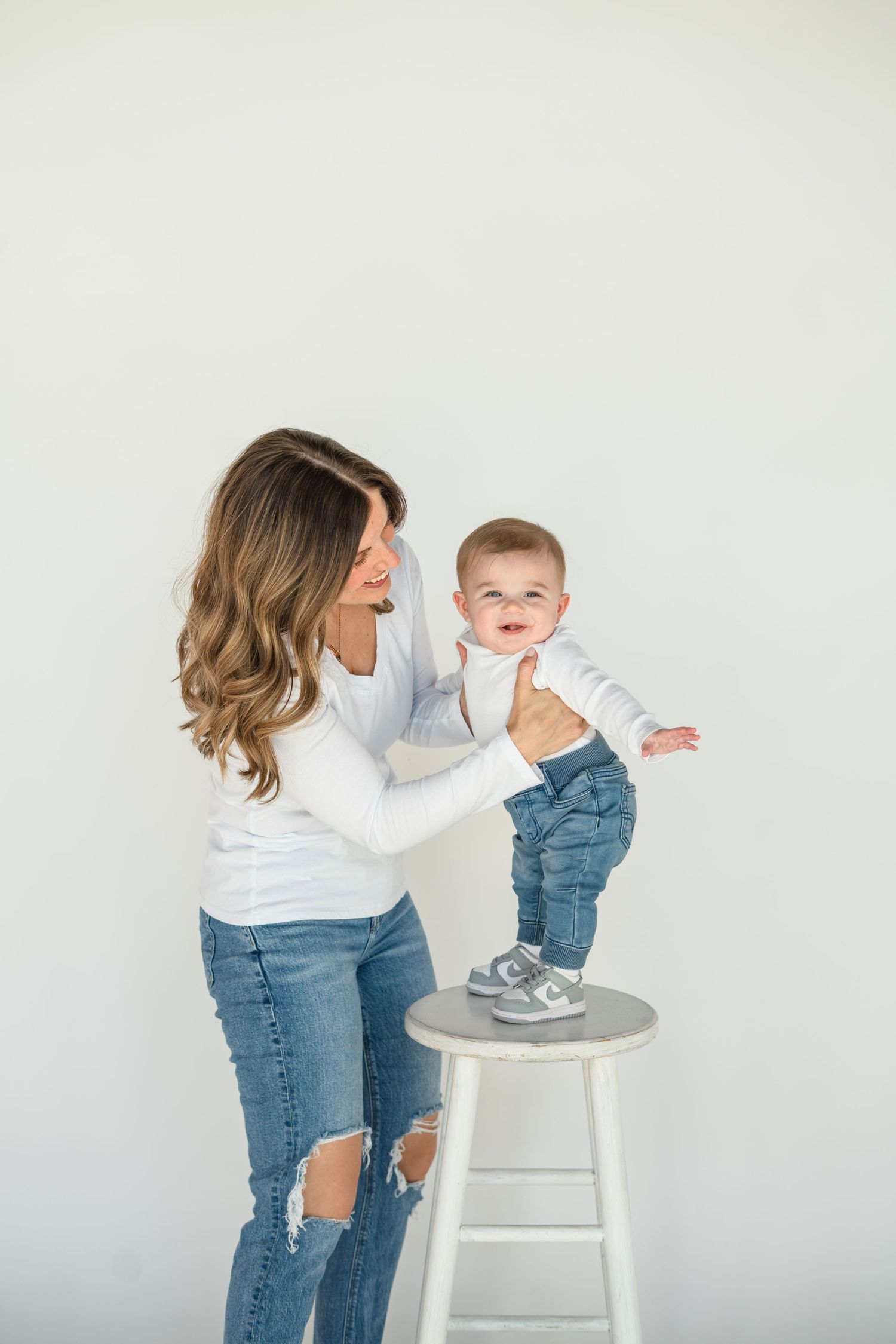 A smiling mother in white top and ripped jeans holds her happy baby boy standing on a white stool in a studio.