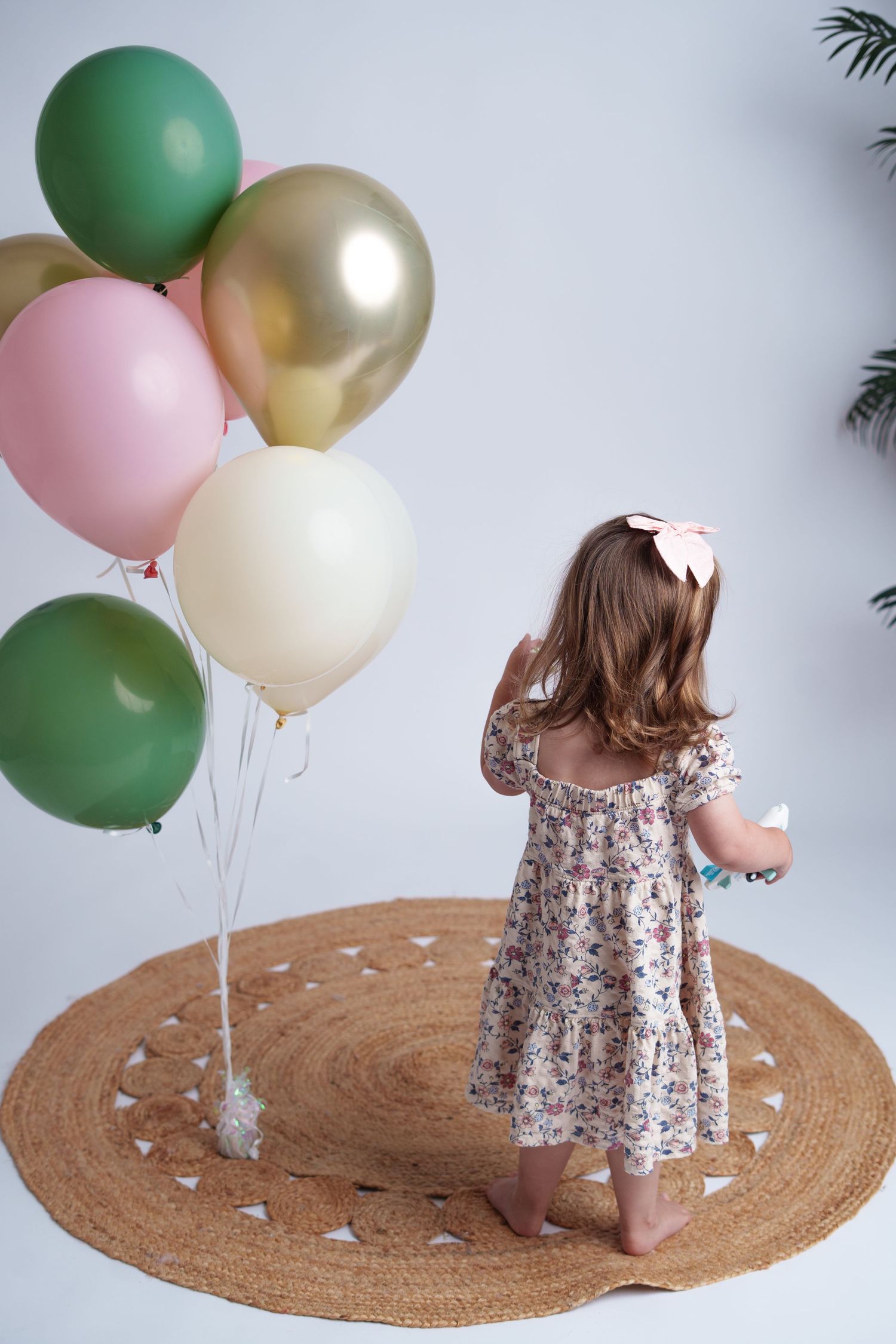 Young girl in floral dress standing on round jute rug with colorful balloons in green, gold, pink, and cream.